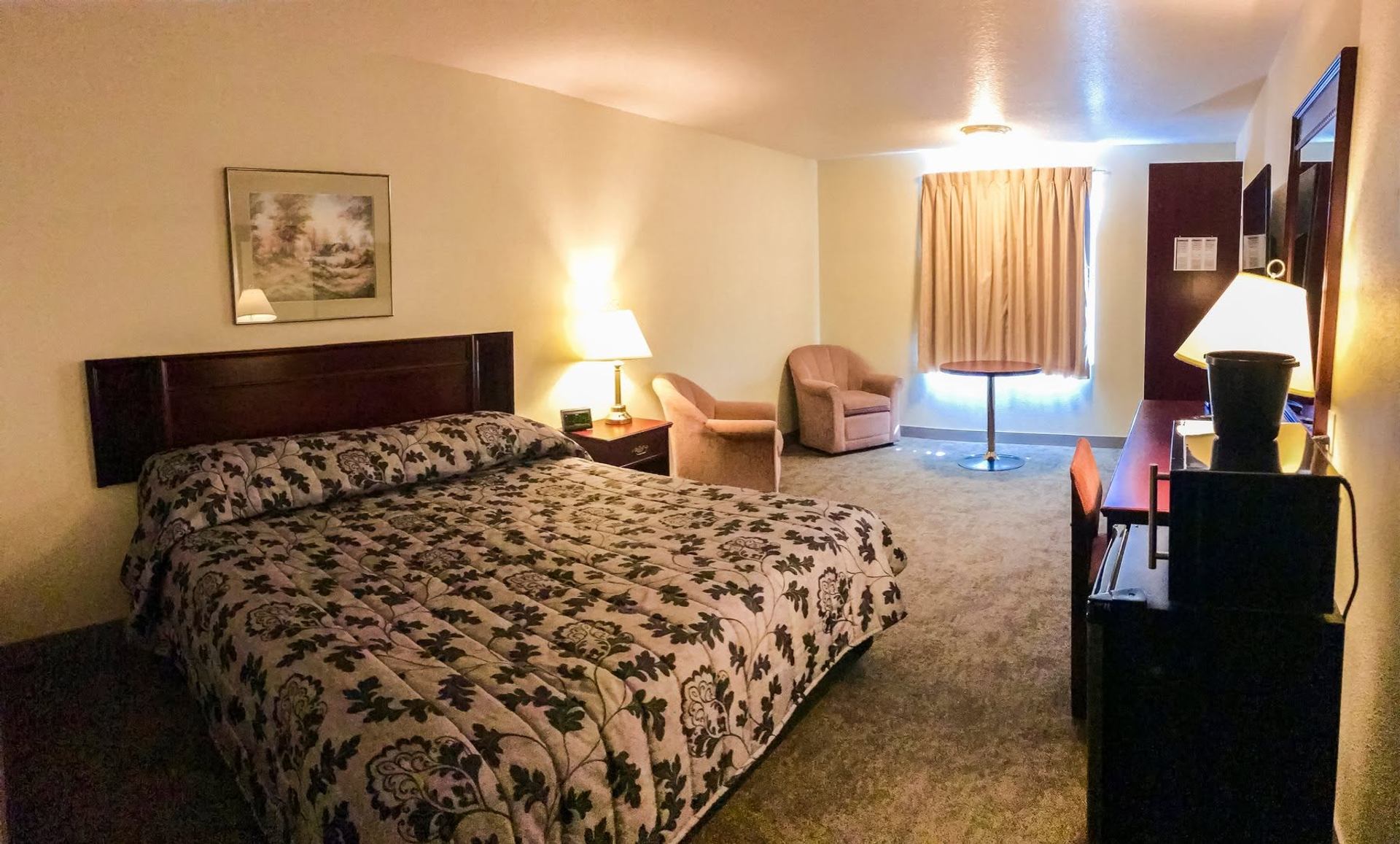 Hotel room with patterned bedspread, armchairs, table, desk, lamps and curtained window.