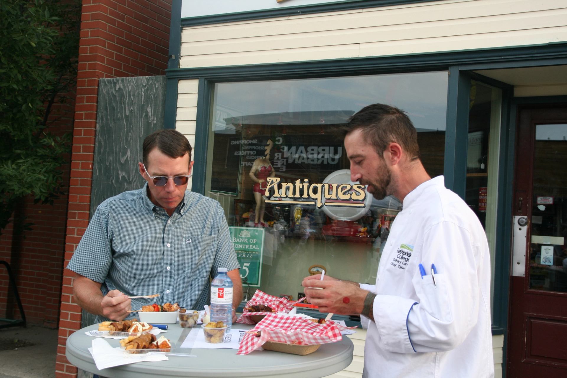 Two men eat at a table in front of an antique store.