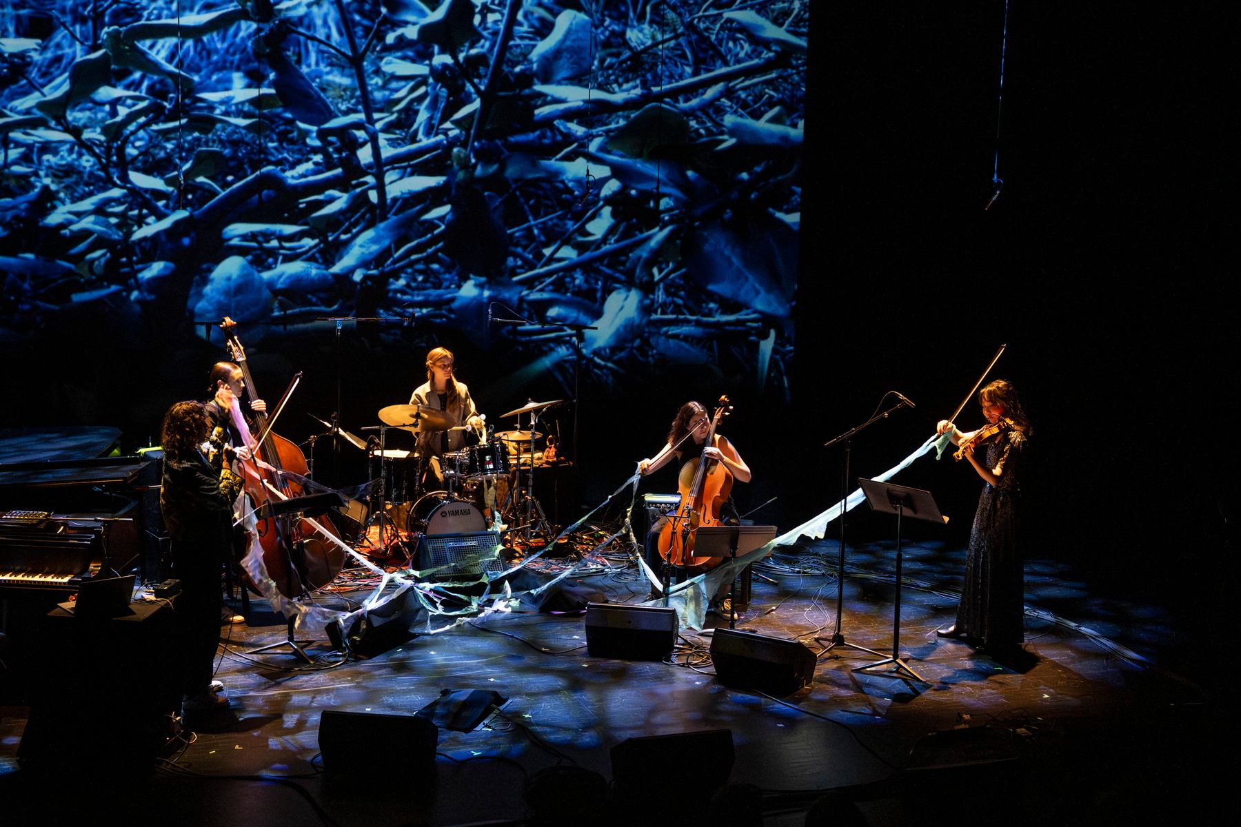Musicians perform on stage with strings, drums and keyboards before a blue abstract backdrop.