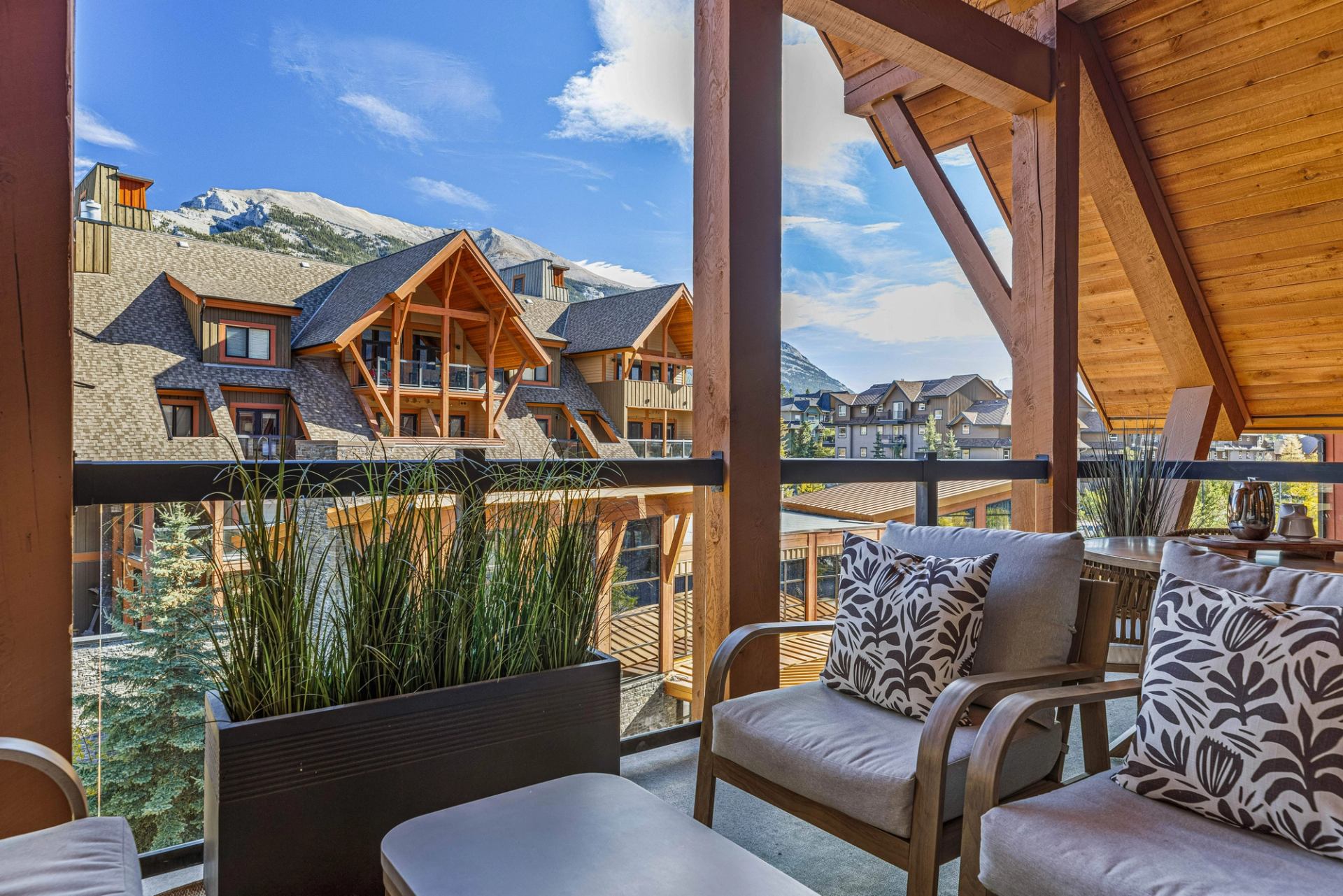 Balcony seating area overlooking timber lodge buildings with mountain backdrop under blue sky.