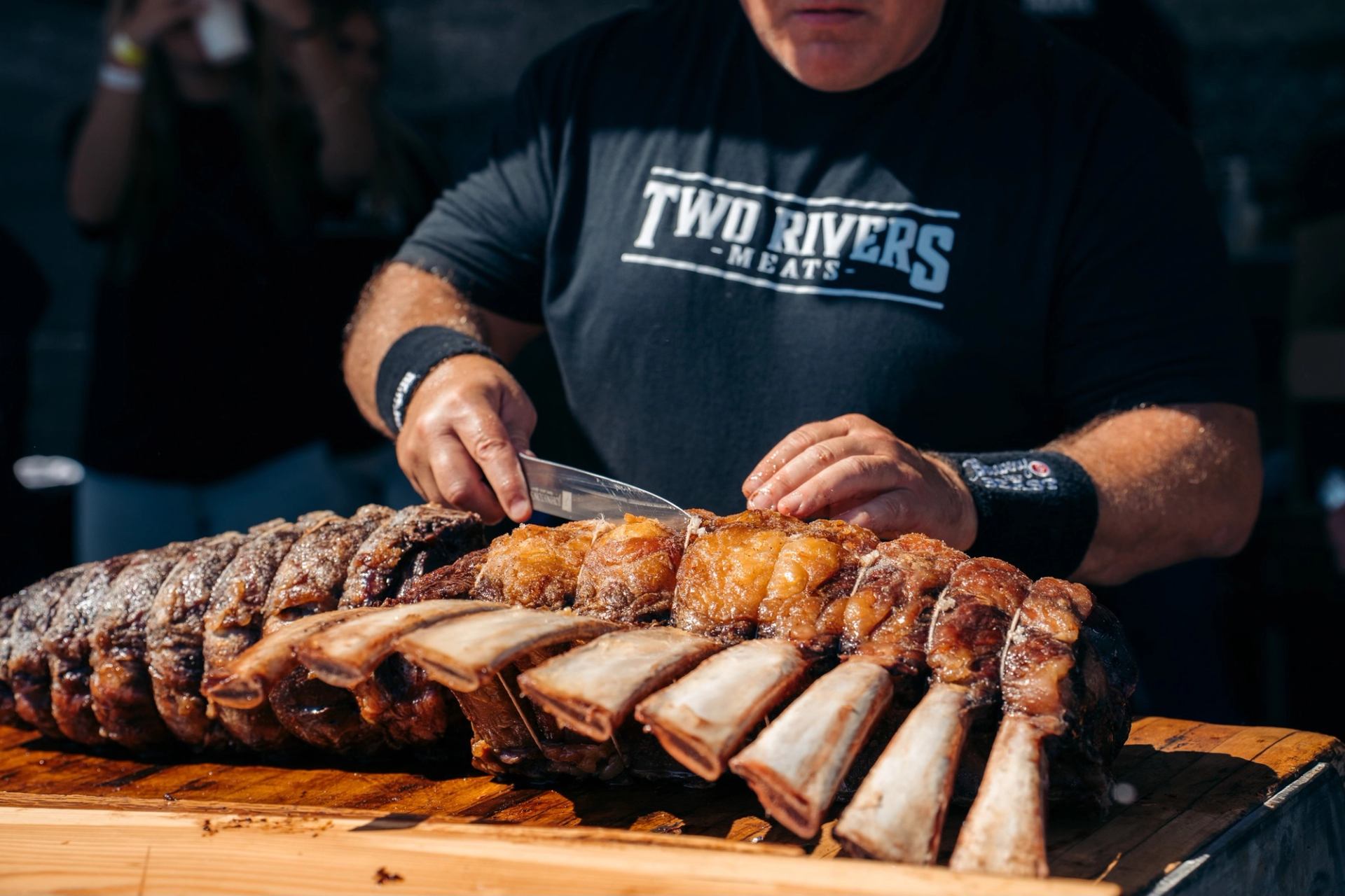 Cook slicing large rack of roasted beef ribs on wooden table outdoors.