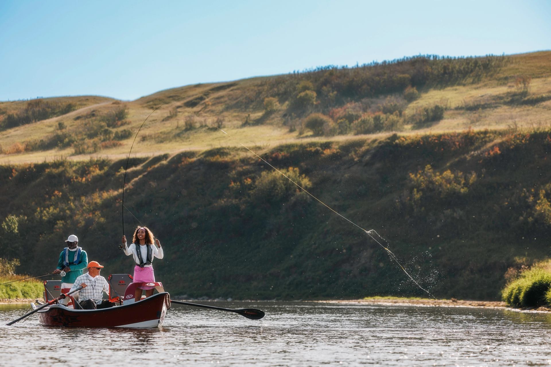 A woman fly fishing from a boat on a river, her line splashing water, with two other people and rolling hills in the background.