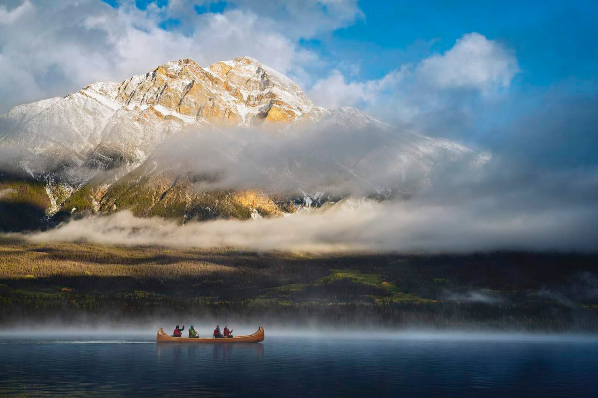 Canoe with paddlers moves across misty lake below snow-dusted mountain and low clouds