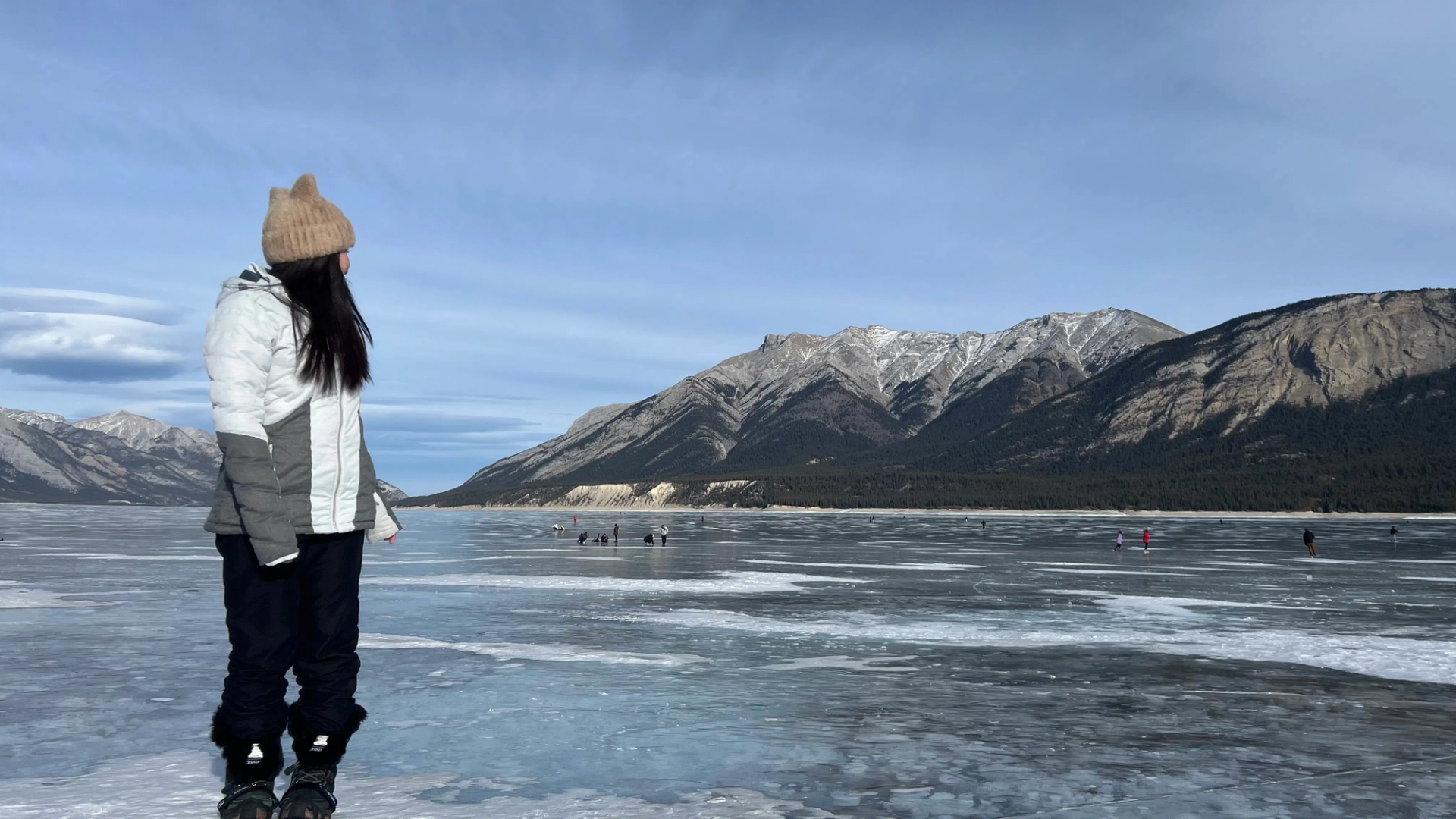 Person in winter clothing stands on frozen lake with distant skaters and mountain backdrop.