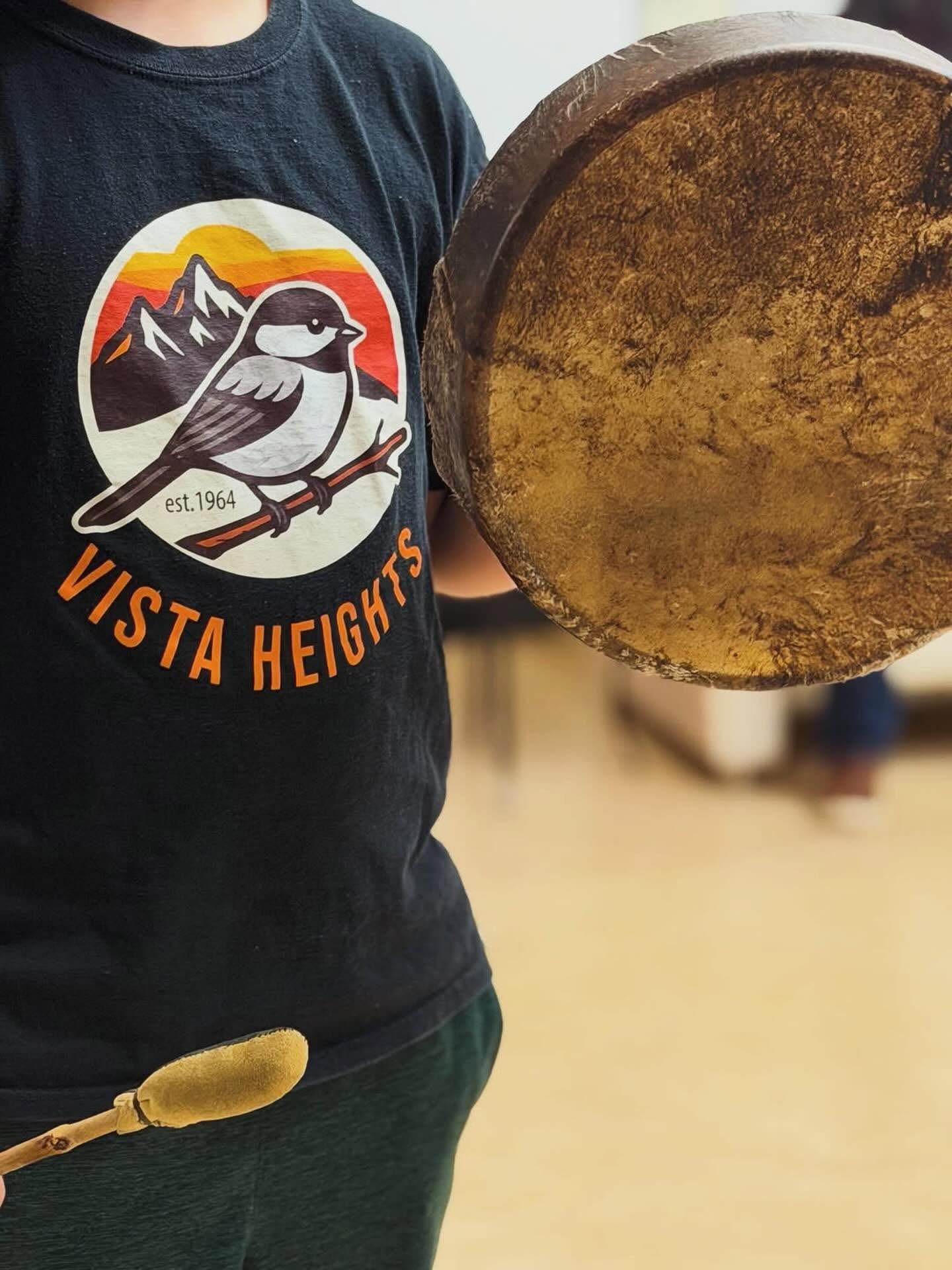 Person in Vista Heights bird T-shirt holding hand drum and padded drumstick.