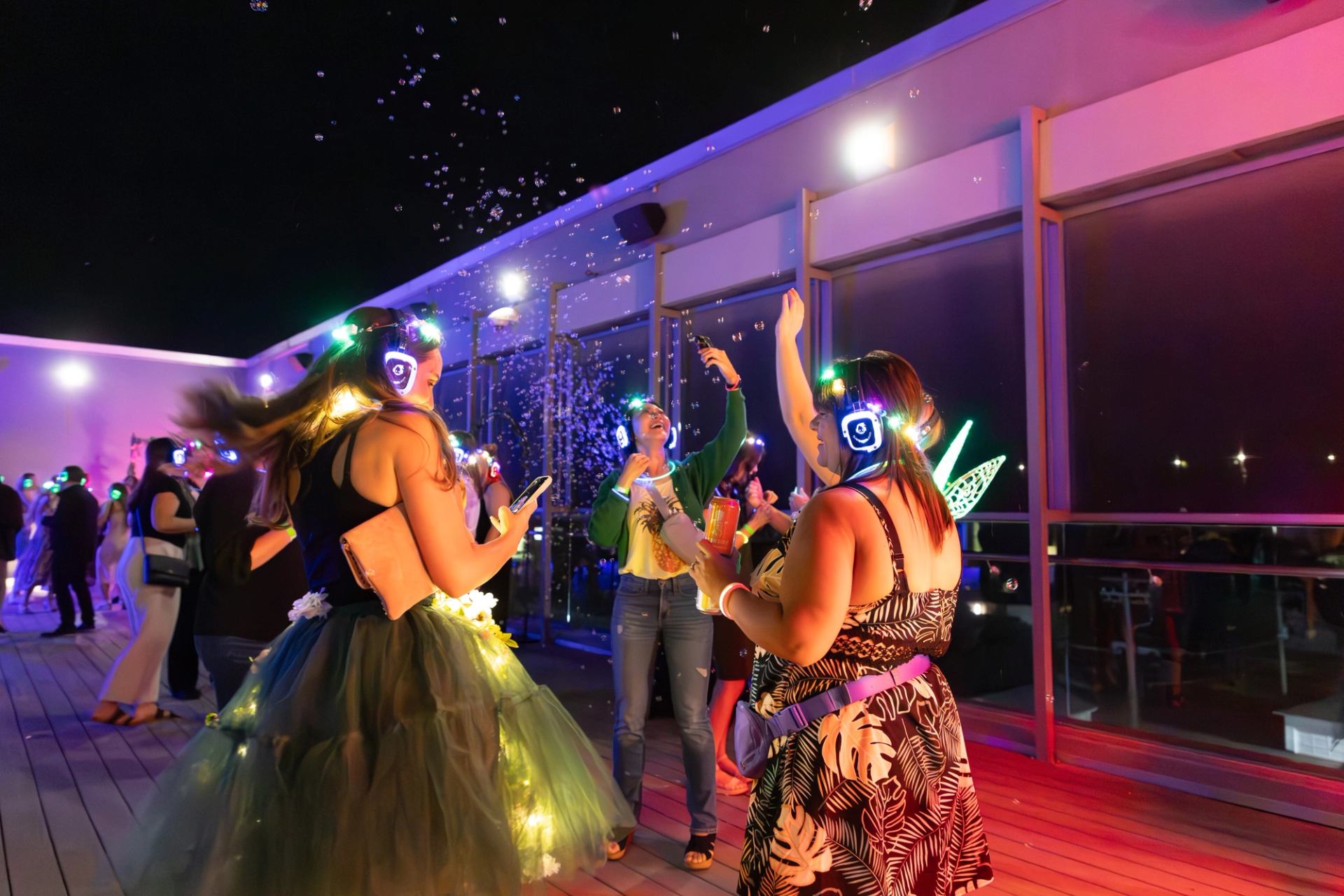 People wearing glowing headphones dance on a rooftop deck surrounded by bubbles at night.