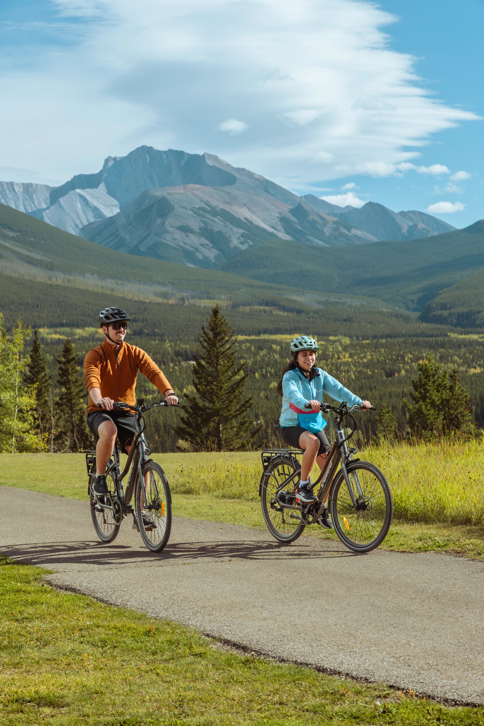 Cyclists wearing helmets ride along a paved path with forested mountains behind.