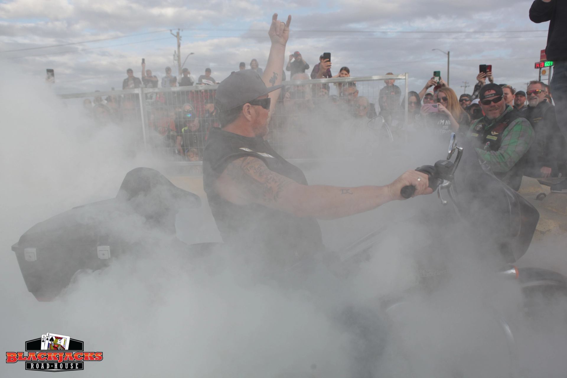 Motorcycle racer waves from smoky burnout competition area; spectators film from behind fencing.
