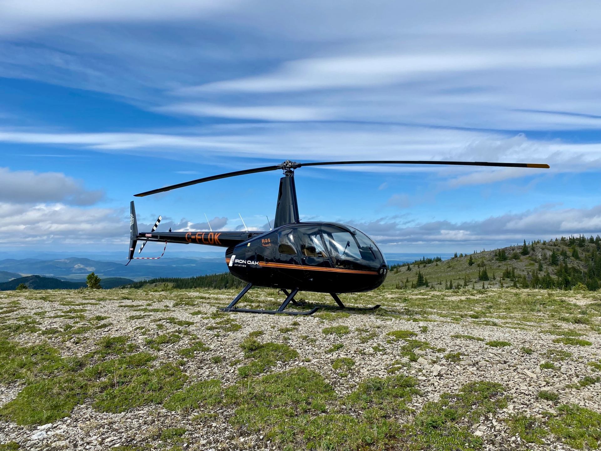 Black helicopter parked on rocky hilltop with distant forested mountains and blue sky.