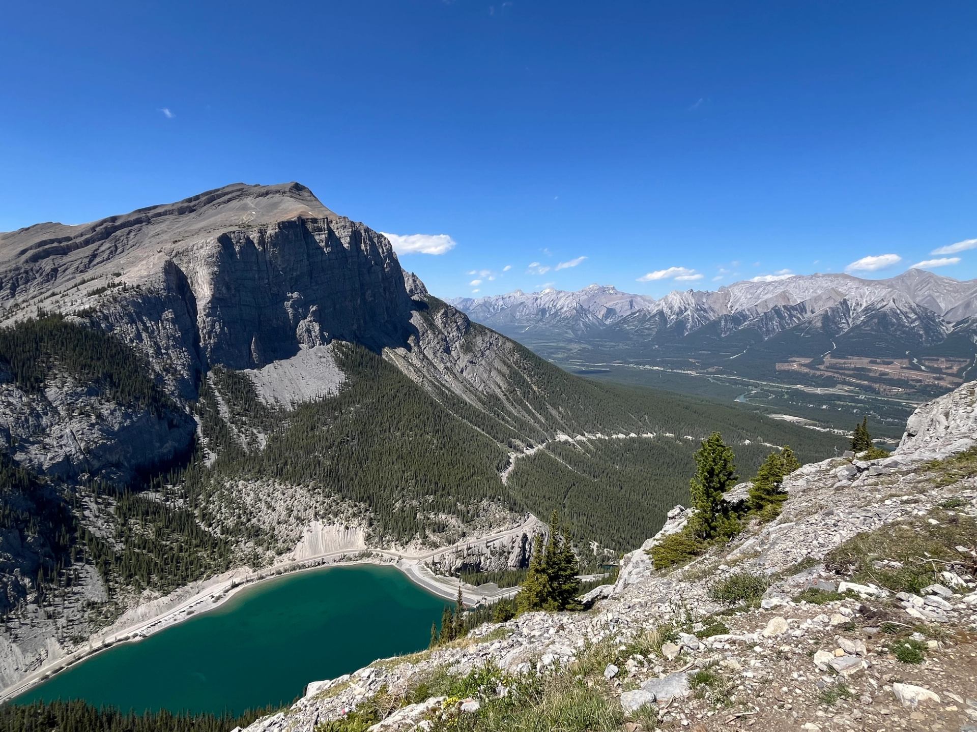 Rocky mountain ridge above turquoise lake and forested valley under clear blue sky.