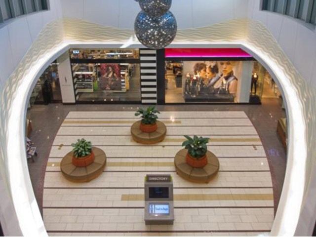 Shopping mall atrium with circular planters, benches, hanging light fixture and storefronts.
