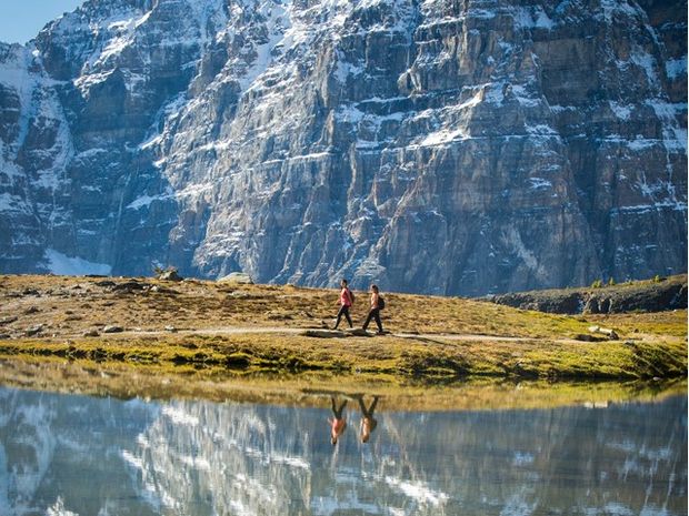 Hikers on a trail with views of mountain peaks reflecting in an alpine lake at Sentinel Pass.