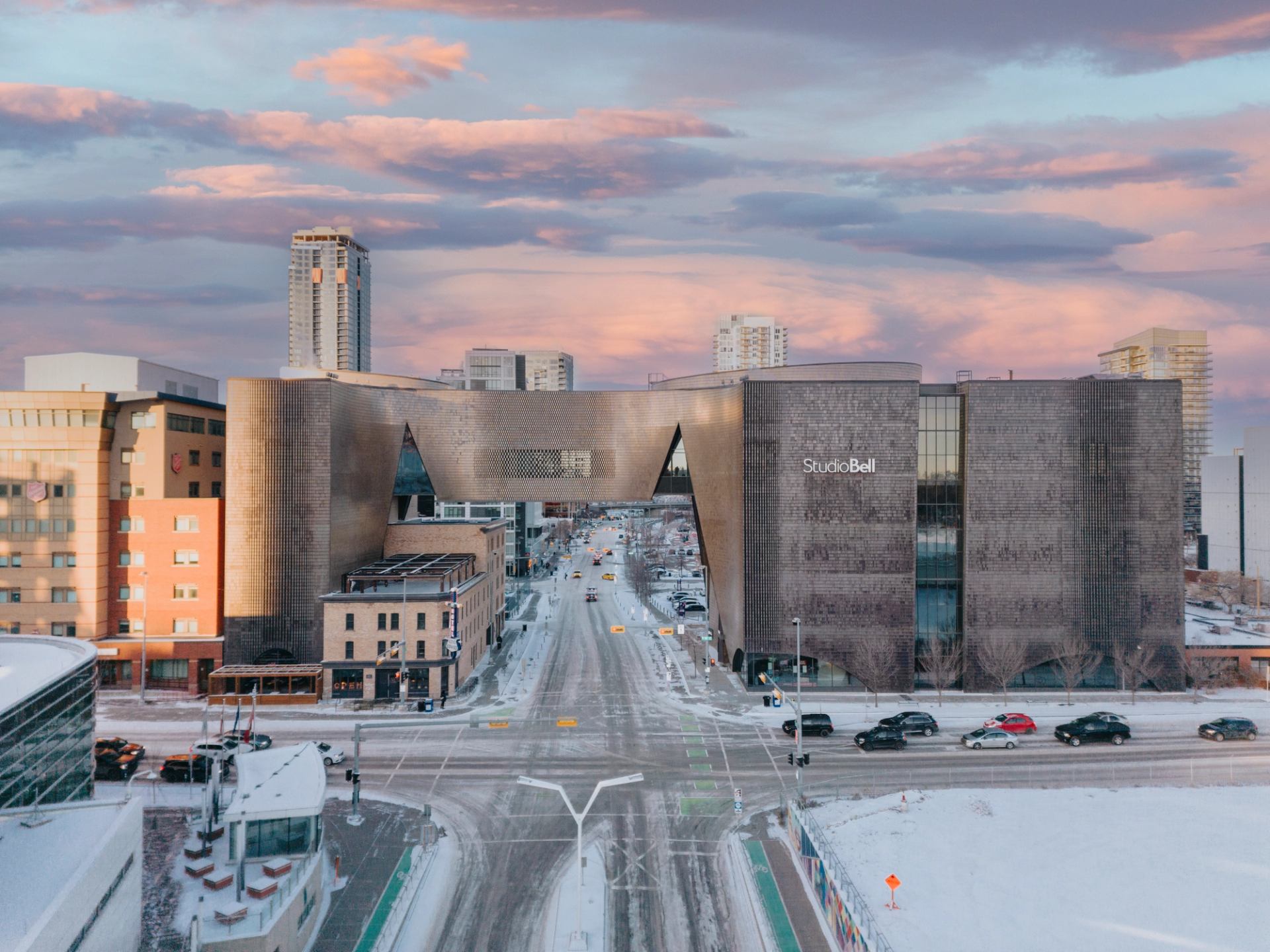 Studio Bell building spans over snowy downtown street with cars and surrounding high-rises.