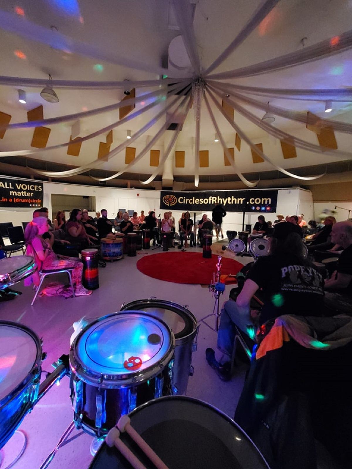 Indoor drumming circle with hand drums arranged around a central rug, streamers overhead, and participants seated in a circle.