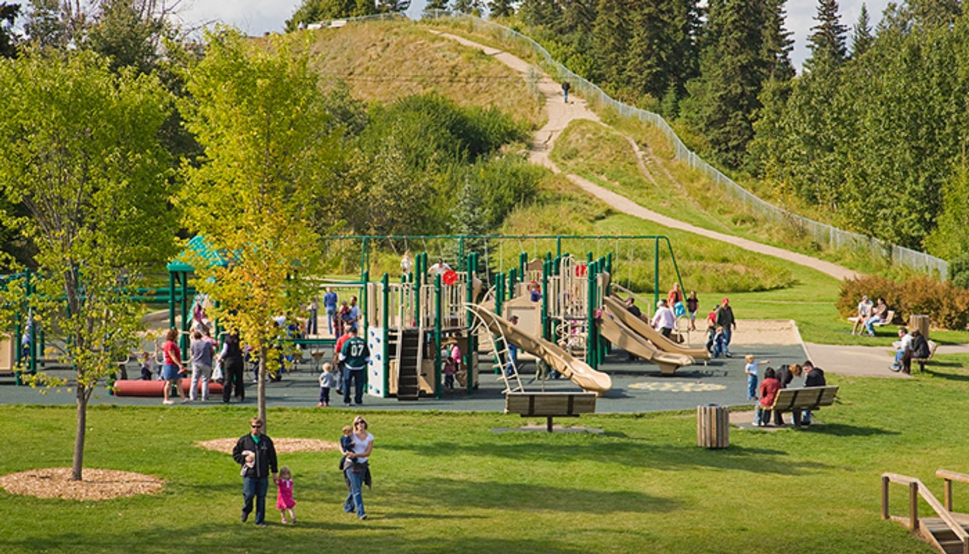 Playground with slides and climbing structures, families walking and sitting on benches in park.