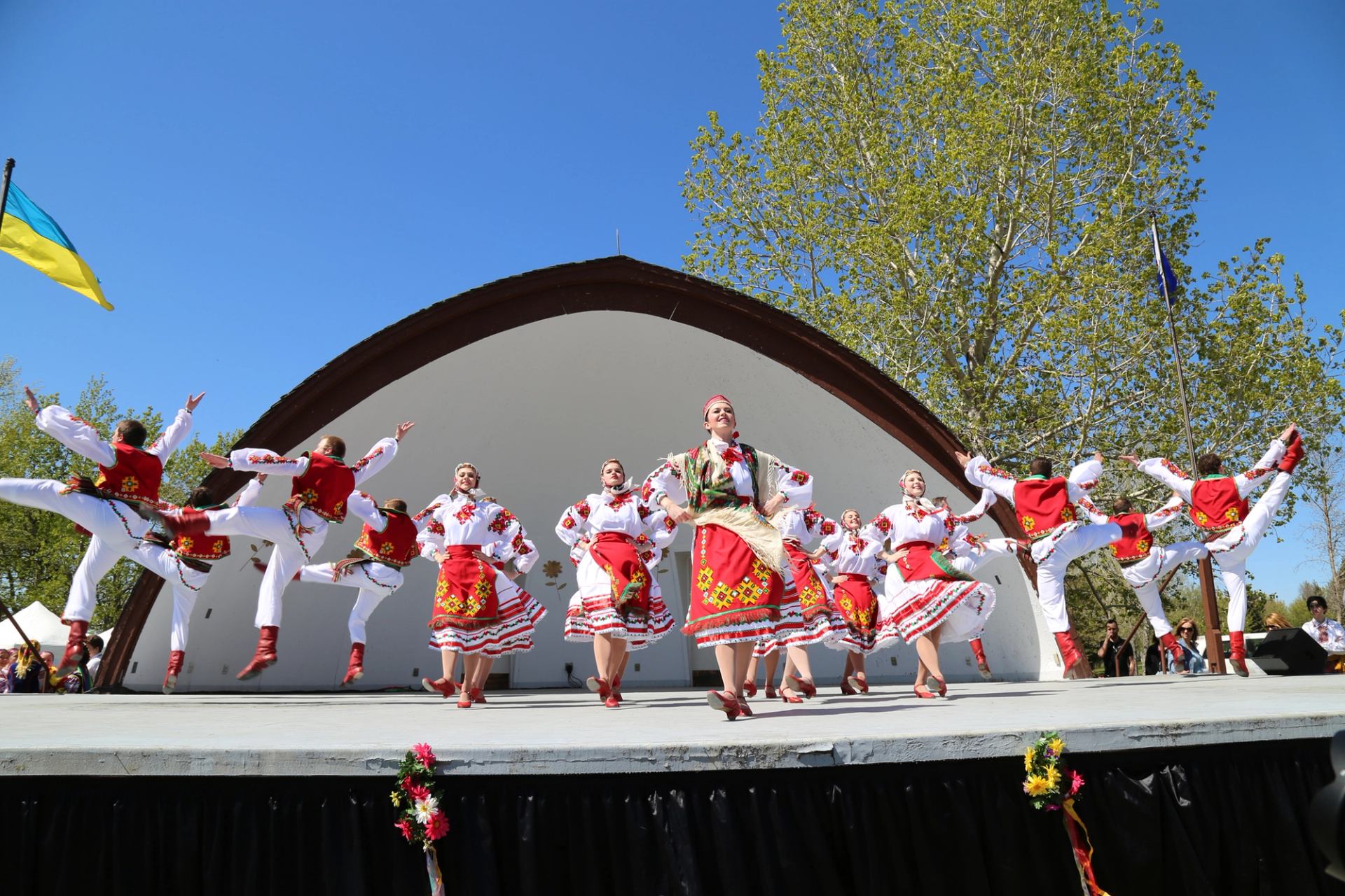 Group of Ukrainian folk dancers performing energetic leaps on an outdoor stage beneath a clear blue sky.