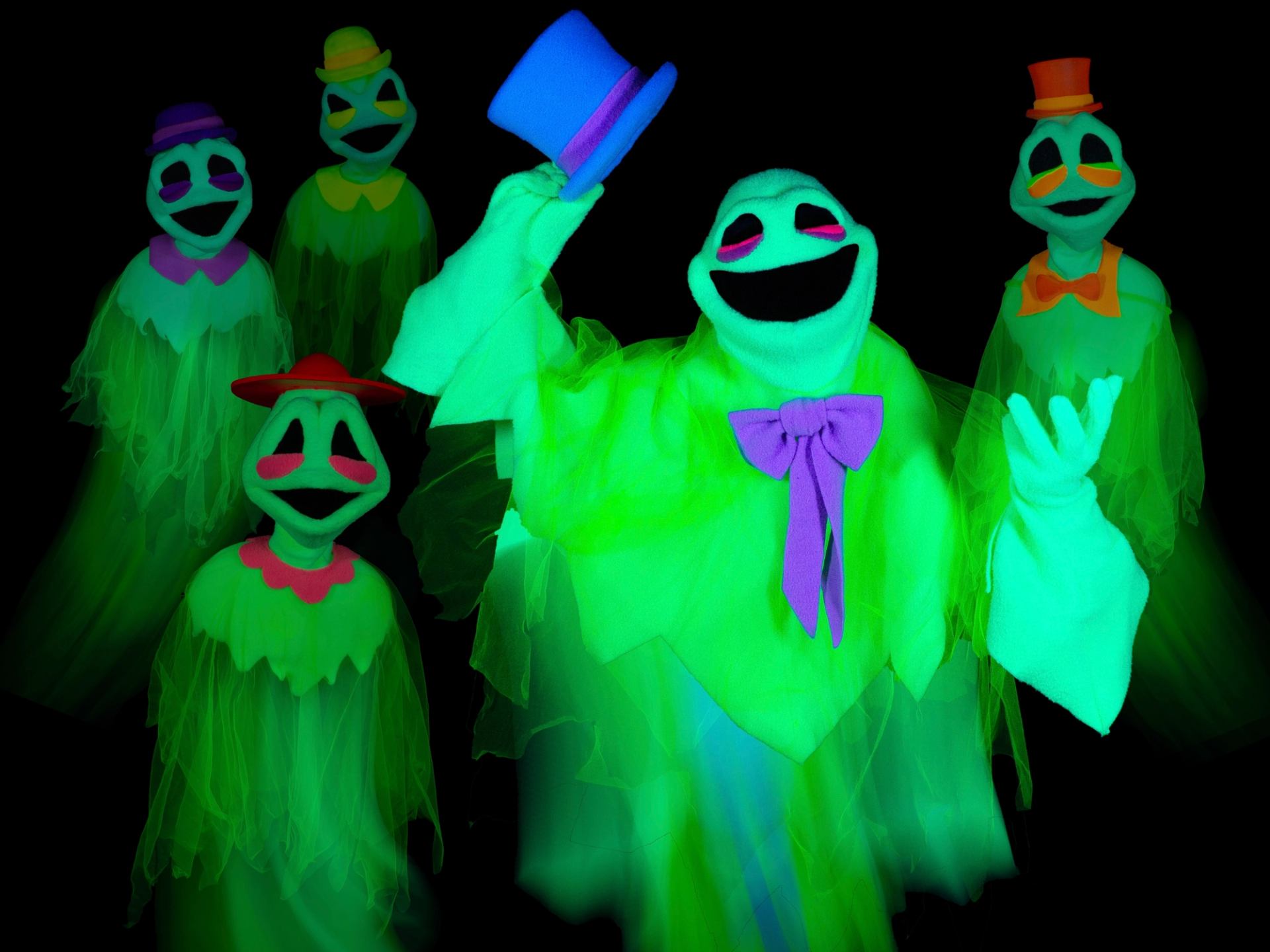 Glowing green ghost puppets in colourful hats and bows pose against a black background.