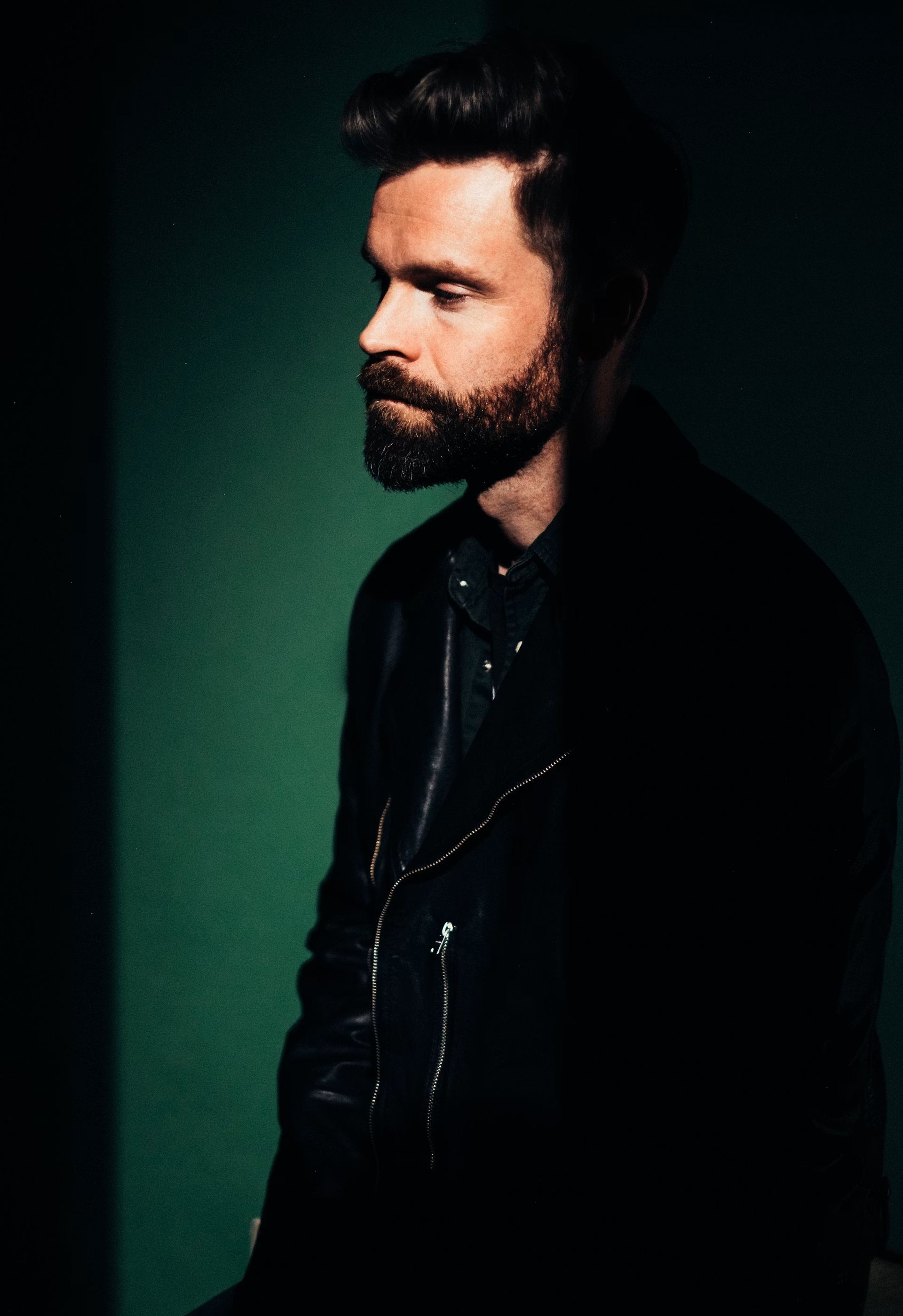 Man portrait with beard wearing a dark jacket against a green studio background.