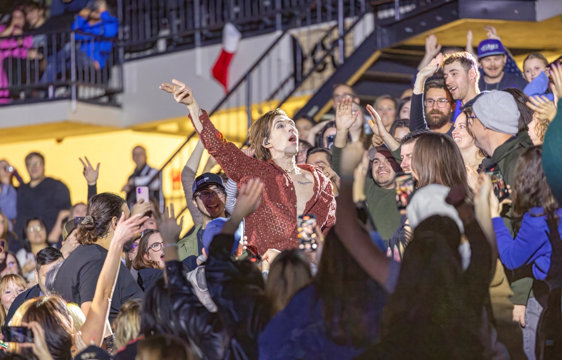 A performer in an open, sparkly red shirt sings to a large, cheering crowd with many hands raised, including people on a balcony.