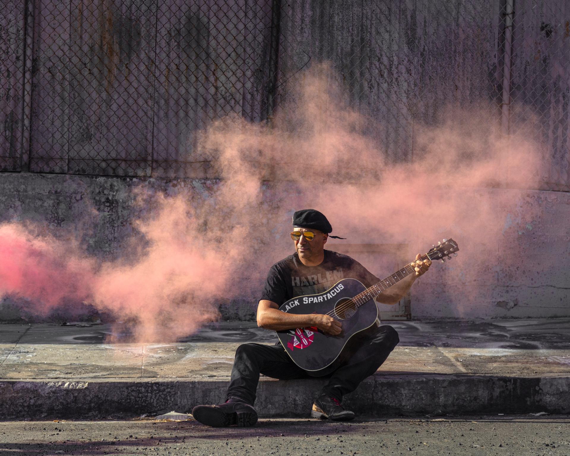 Street musician sits on curb playing guitar as pink smoke drifts behind.