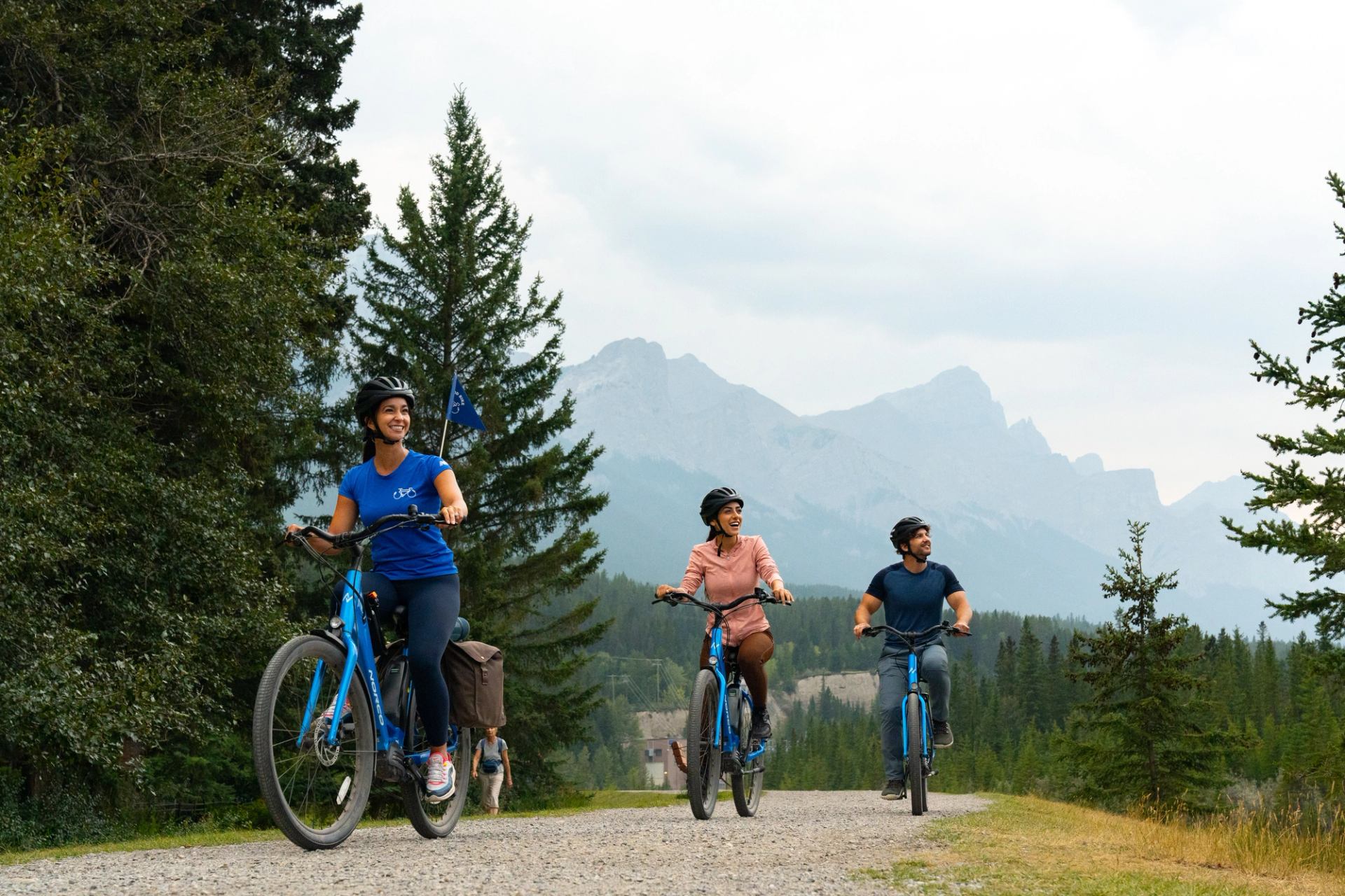 Three smiling people wearing helmets ride blue bicycles on a mountain gravel path.