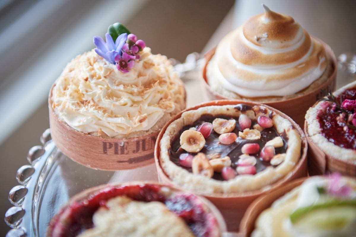 A close-up of a selection of mini pies on a glass tray, including one with meringue, a chocolate tart with hazelnuts and pomegranate, and one with cream and a flower.