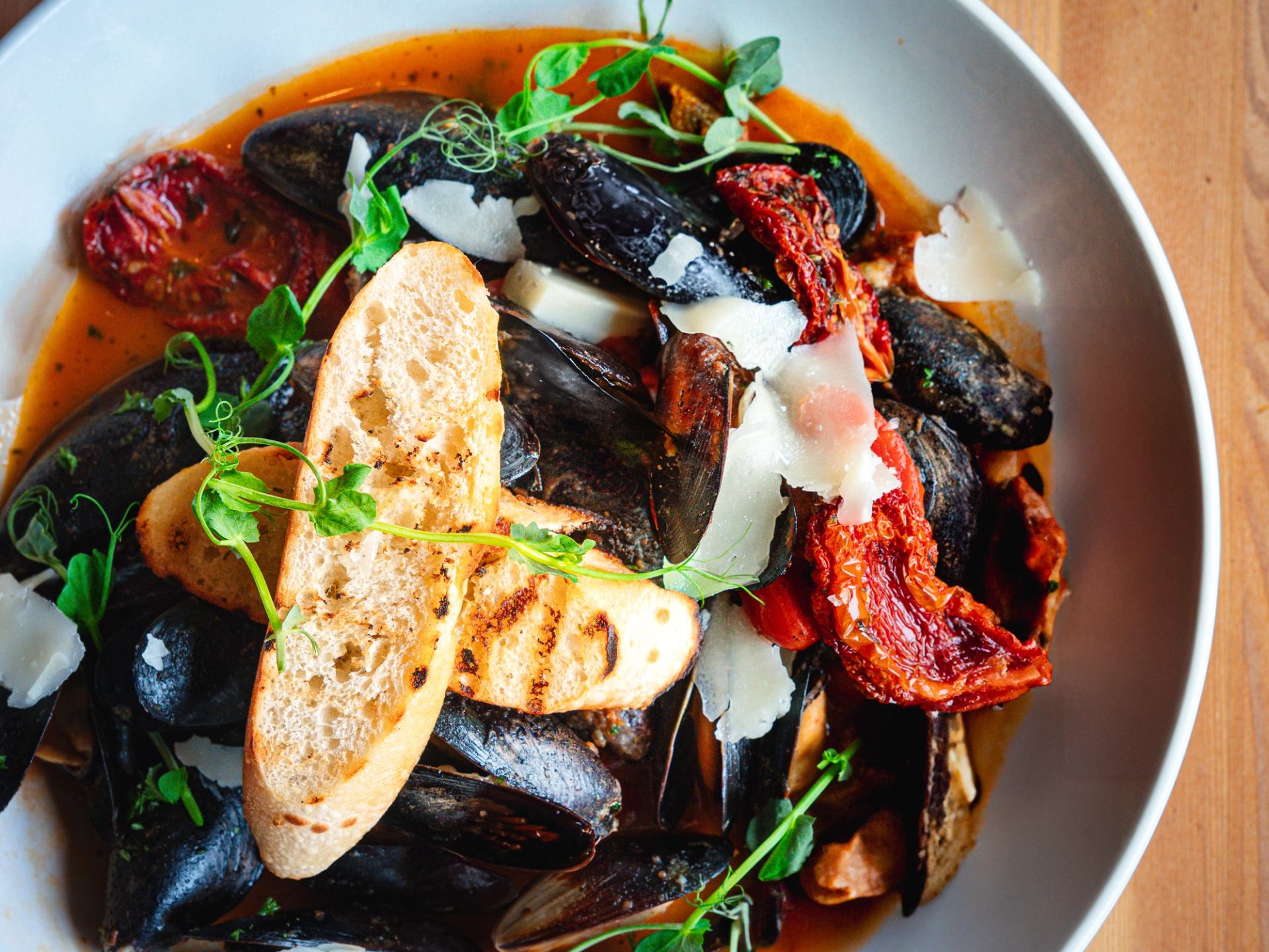 Bowl of mussels in red broth with herbs, cheese, and toasted bread.