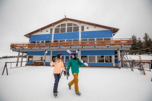 Two skiers with their skis slung over their shoulders, walk and talk at the base of the hill, lodge in background, before skiing Hidden Valley Ski Resort.