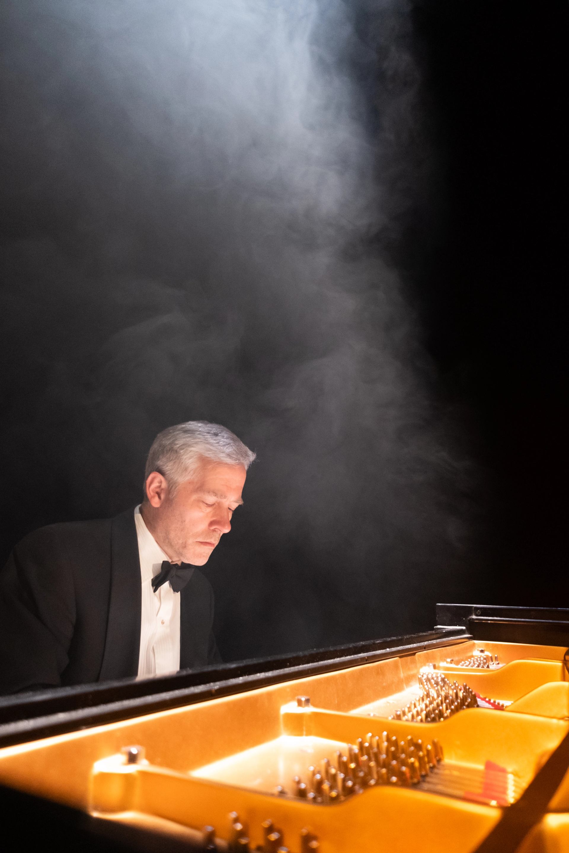 Pianist in tuxedo plays grand piano under stage light with rising smoke.