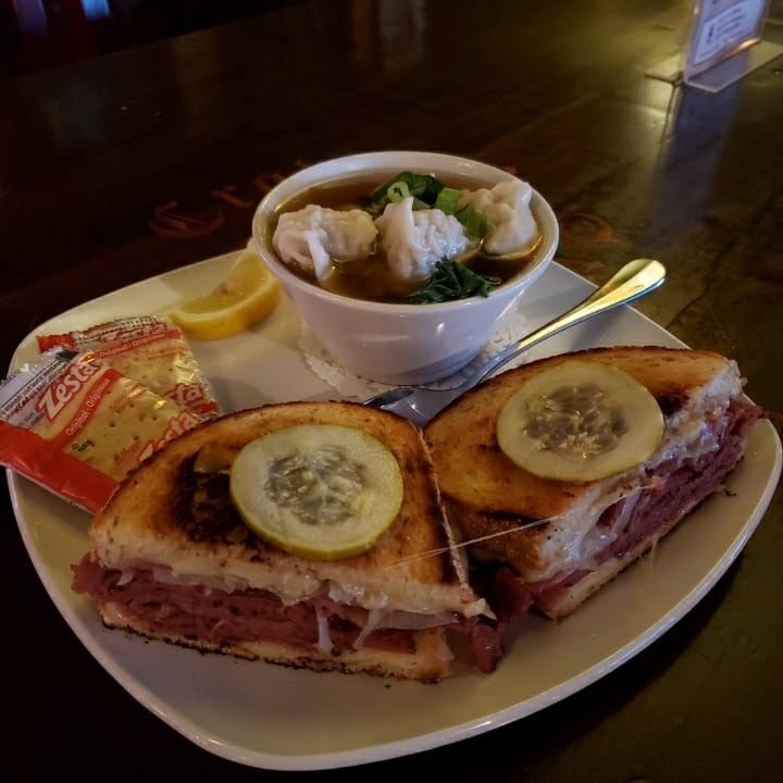 A plate with a grilled sandwich topped with pickle slices, a bowl of soup with dumplings and greens, a lemon wedge, and cracker packets. Cozy and appetizing.