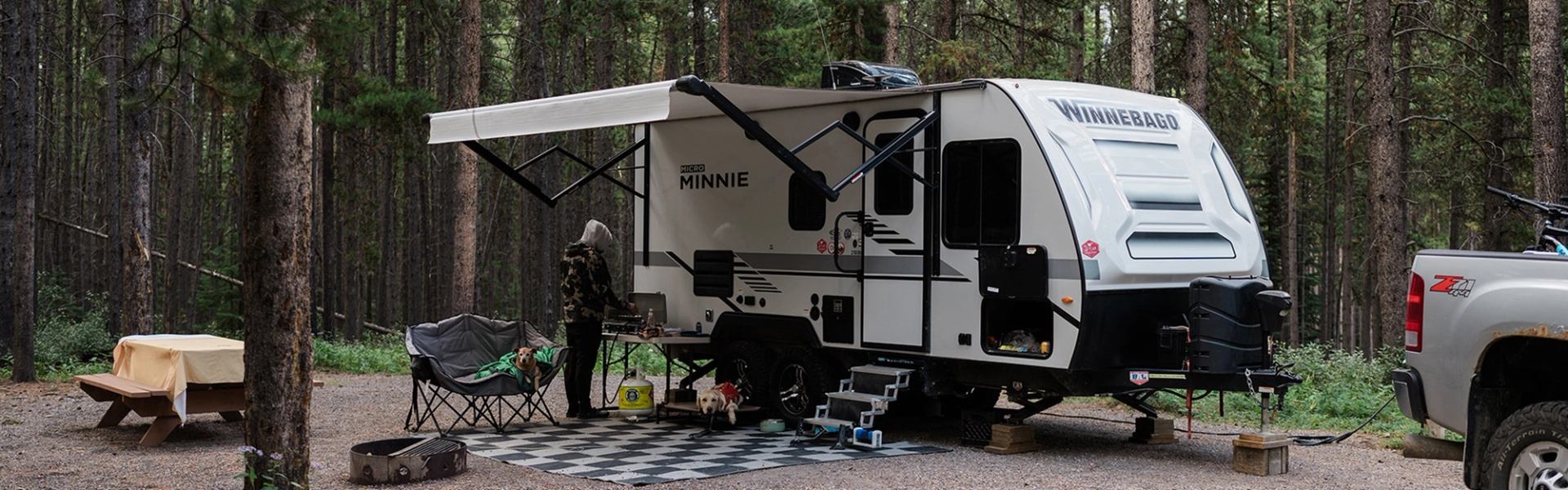 Travel trailer with awning, campsite chairs and table in forested campground.