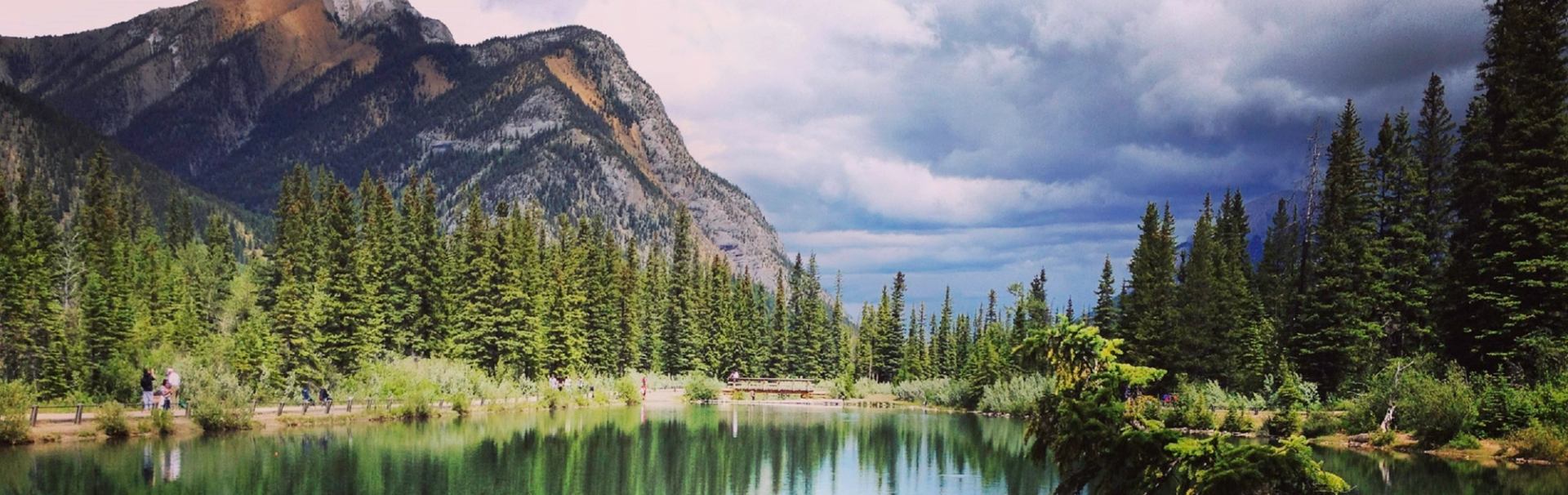 Mountain lake with evergreen forest, walking path and people reflected in calm water