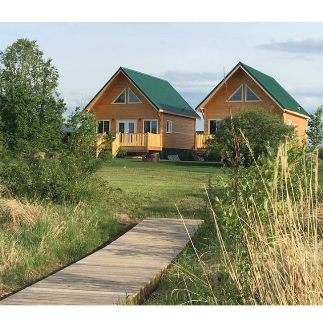 Two wooden cabins with green roofs surrounded by grass and trees, connected by a wooden walkway.