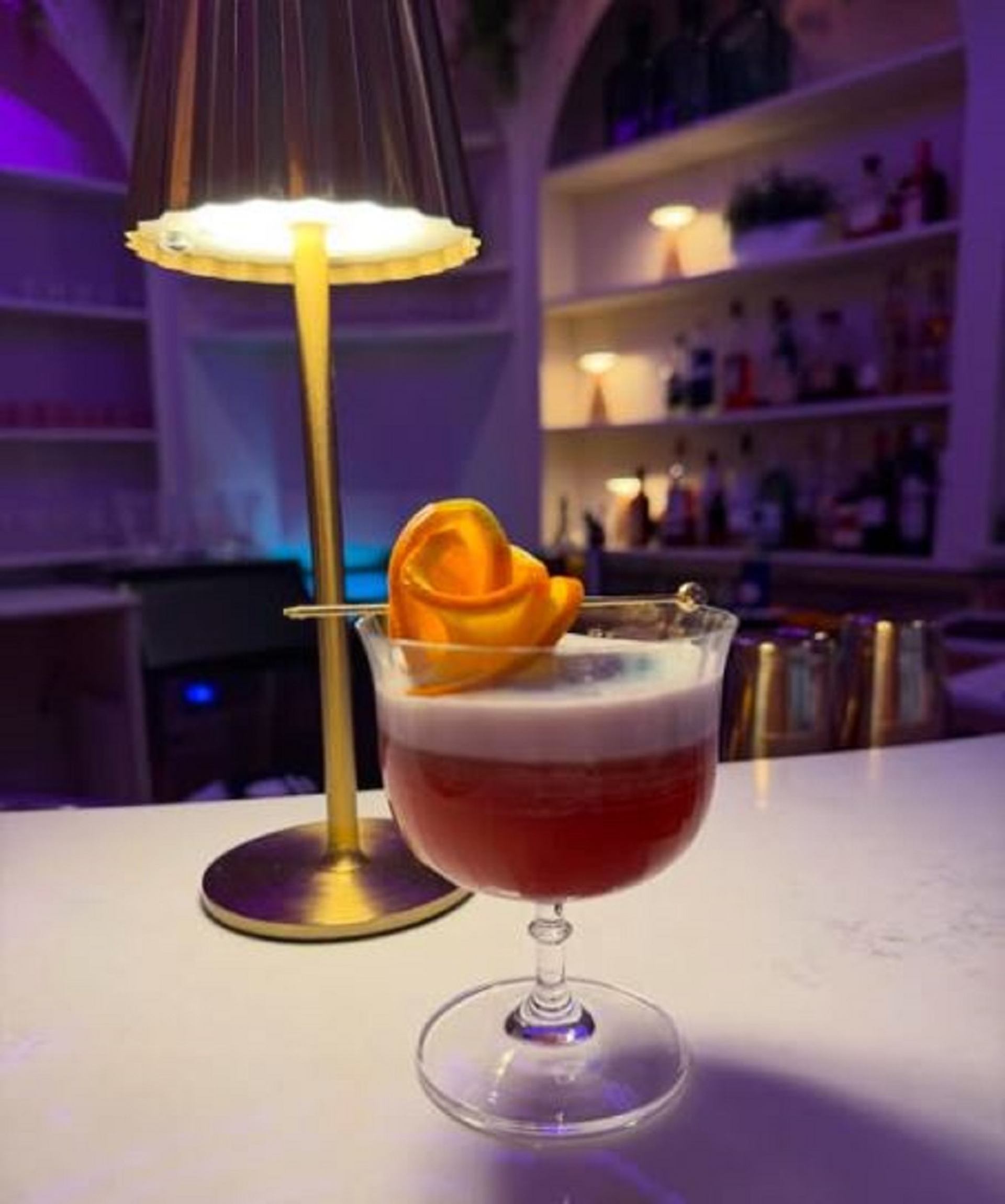 Layered cocktail with citrus garnish beside a small lamp on the bar at Bungalow Club.