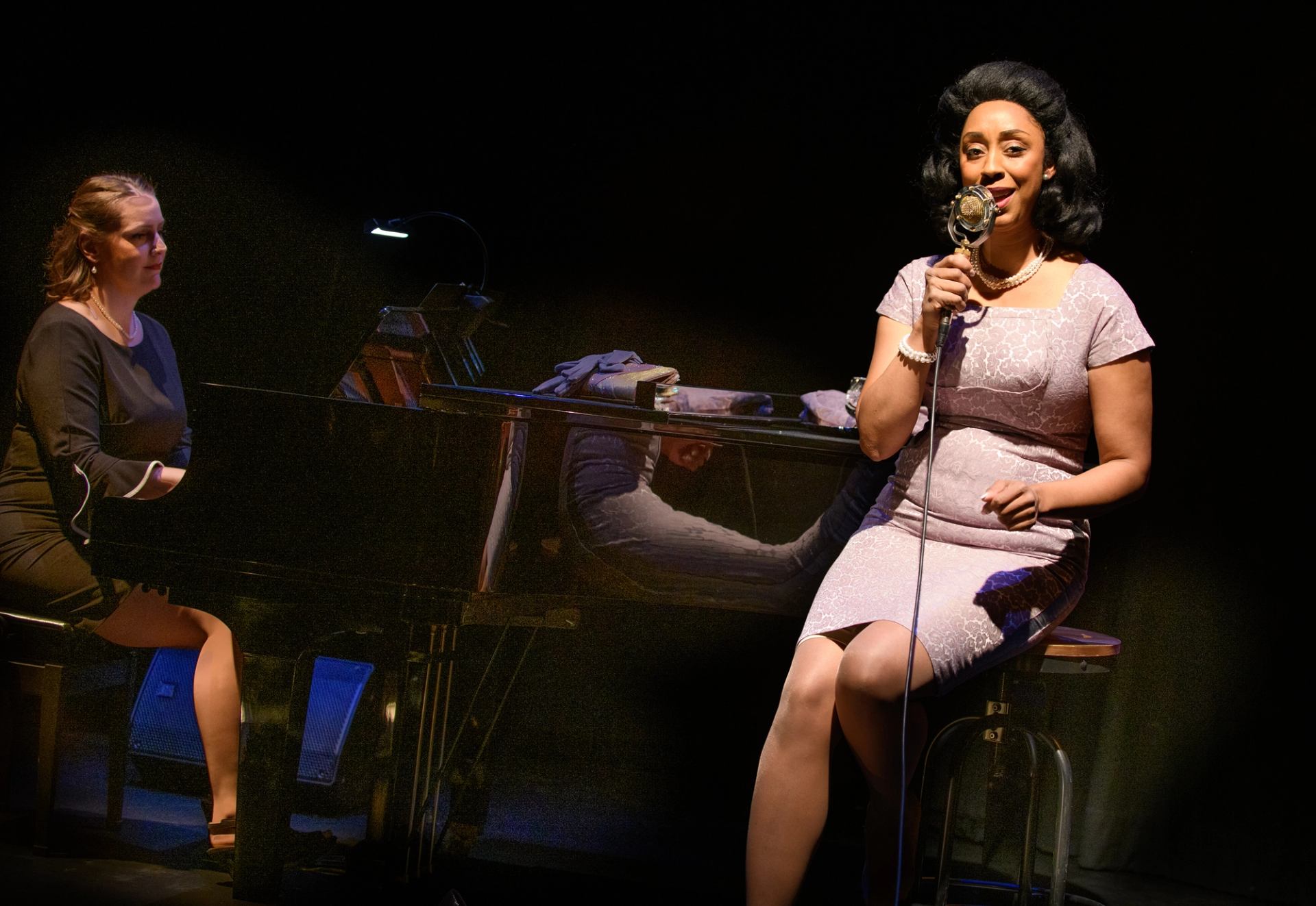 Singer in vintage dress performs into microphone beside pianist at grand piano onstage.
