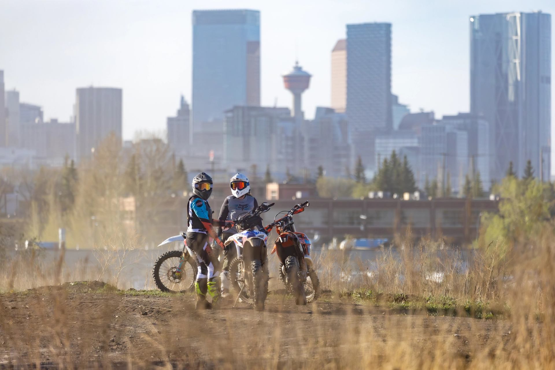 Two riders with dirt bikes on trail overlooking city skyline.