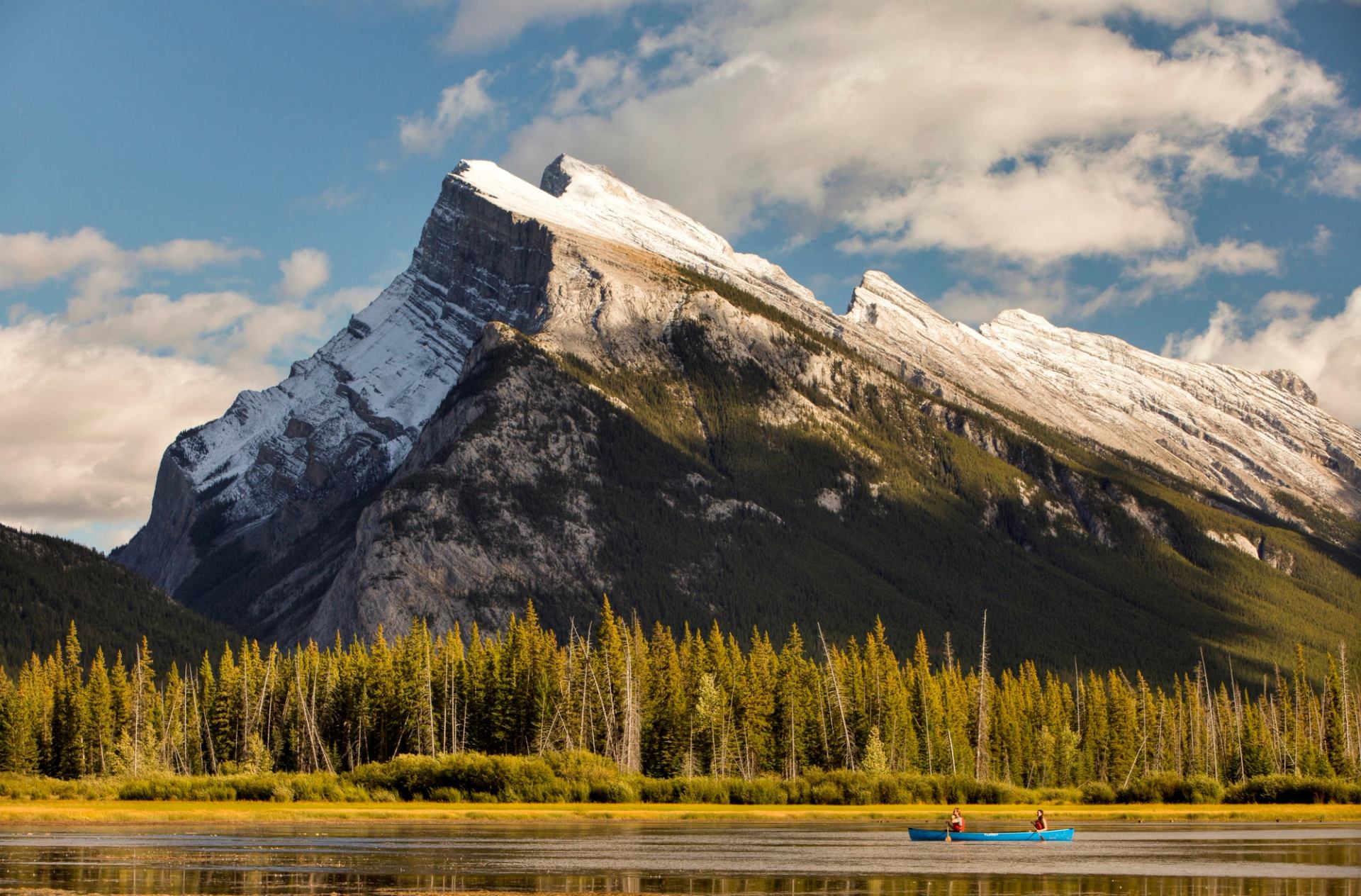Canoeists paddle across a lake below a snow-capped mountain and evergreen forest.