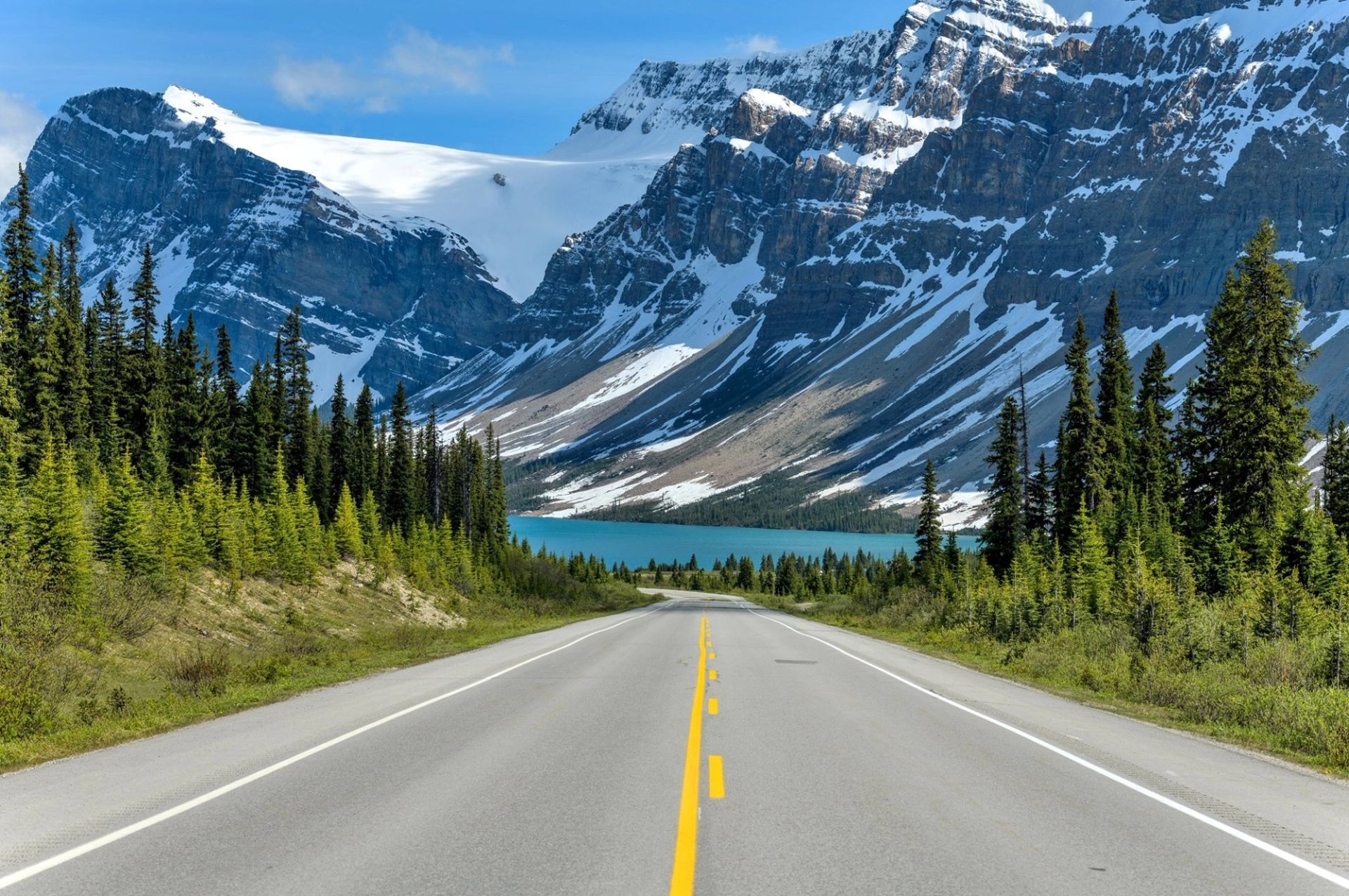 Straight road with yellow lines leading to snow-capped mountains, trees, and water under blue sky.