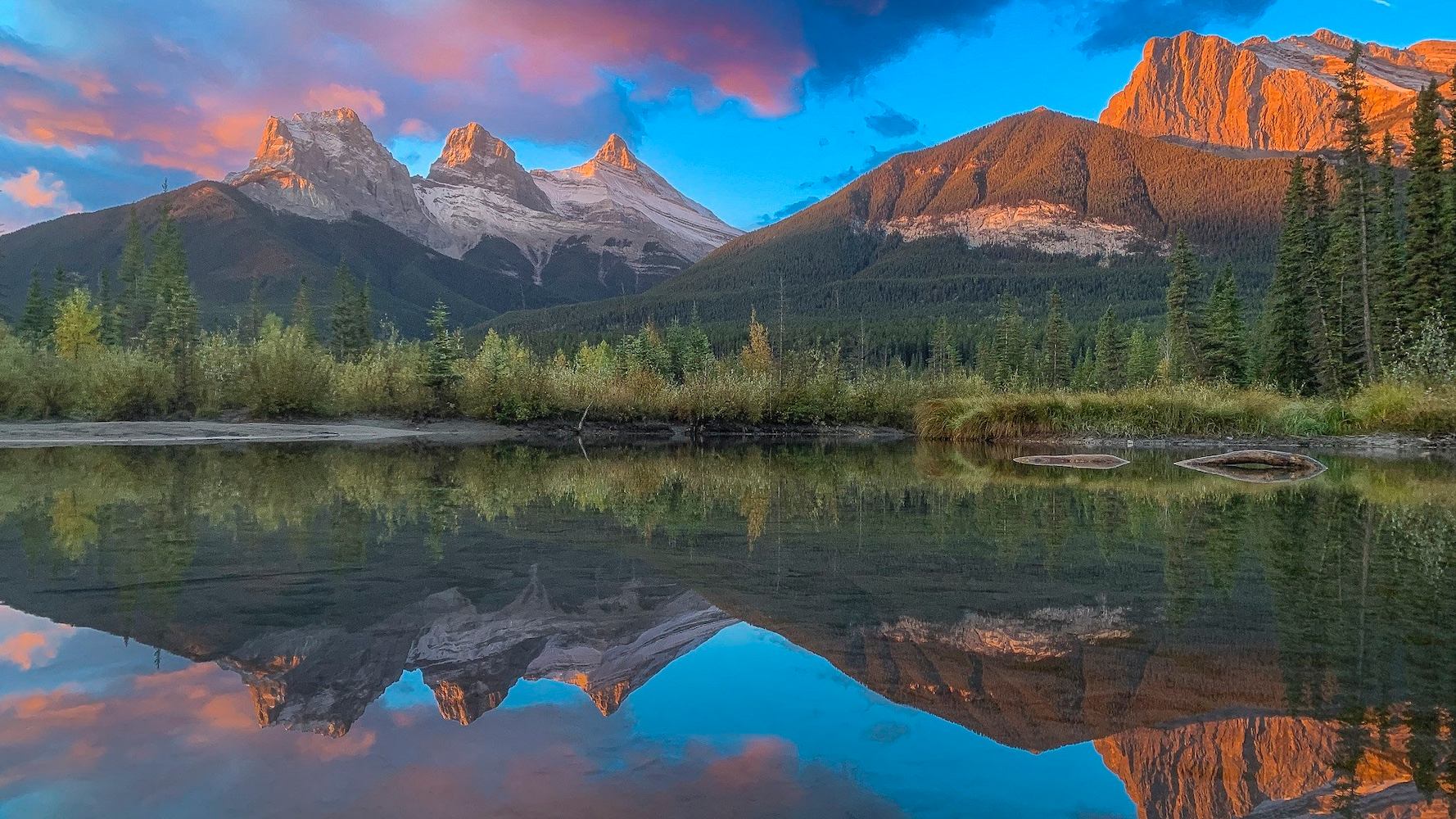 The Three Sisters mountains and a golden-lit peak reflected in still water under a colorful sky.