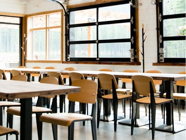 Modern taproom interior with wooden tables, metal chairs, and large sunlit windows.