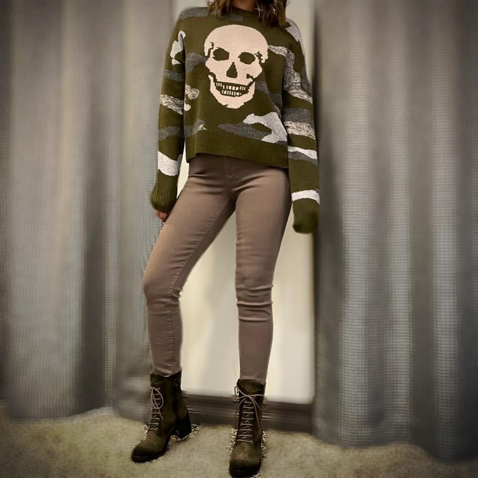 Person wearing olive skull sweater, beige pants, and black lace-up boots against curtains.