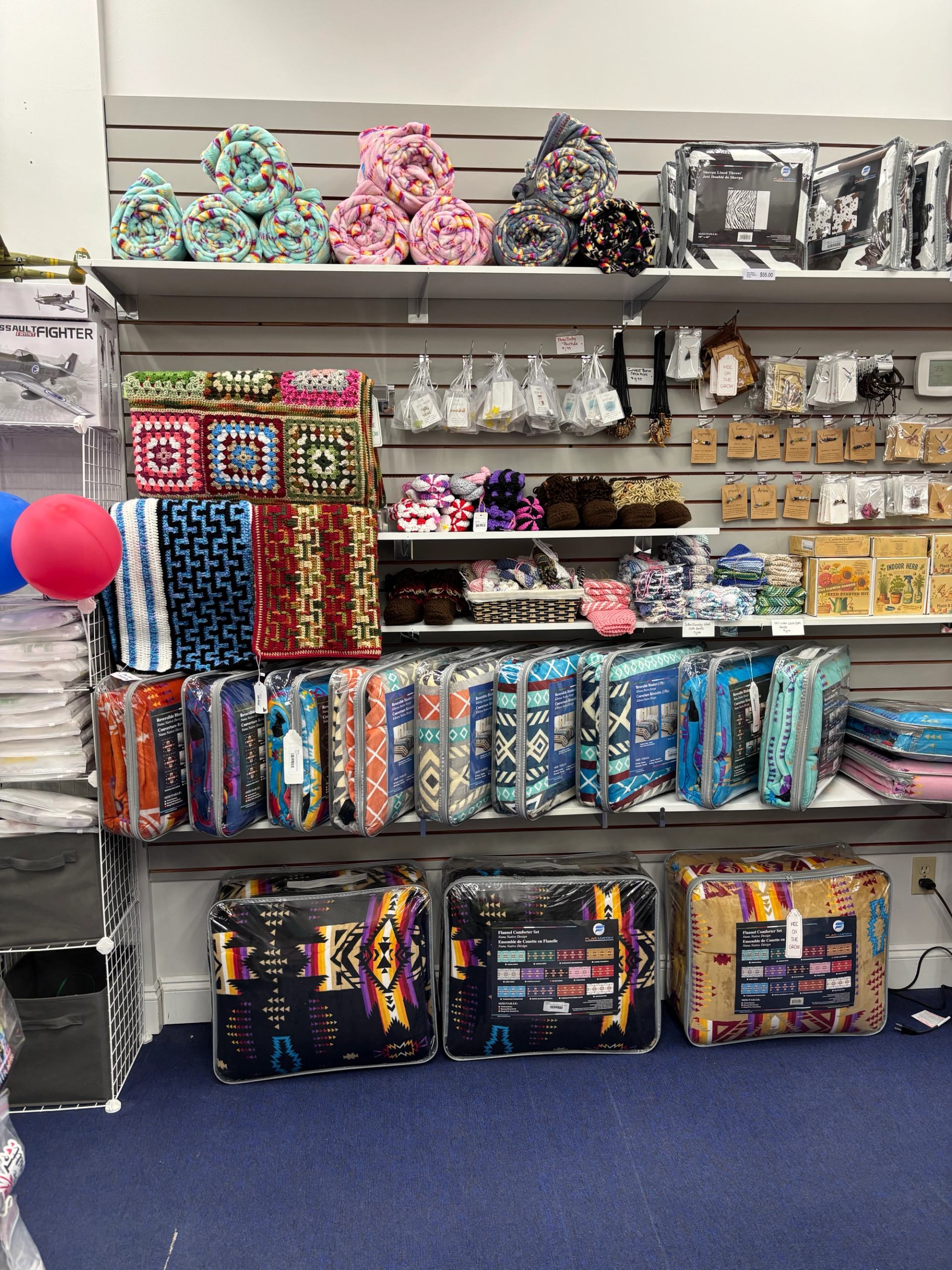 Retail display of folded blankets, patterned throws, handmade crafts, and small gift items arranged on wall shelves.