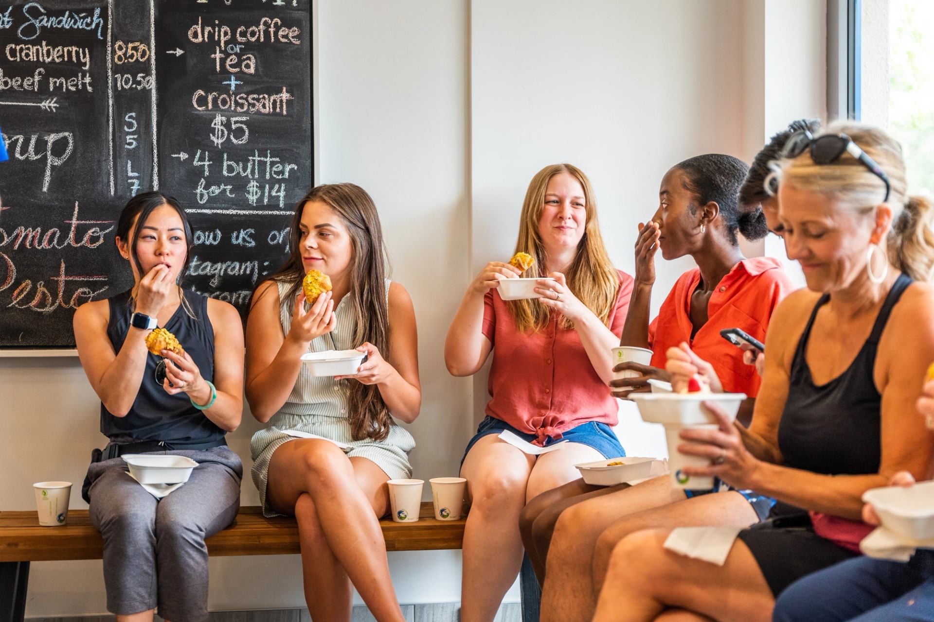 Group seated on bench eating pastries, chalkboard café menu in background.