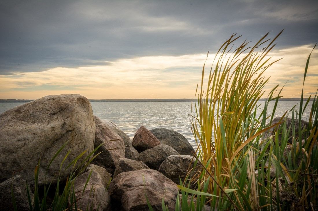 Large shoreline rocks and tall grass beside calm water under cloudy sky.