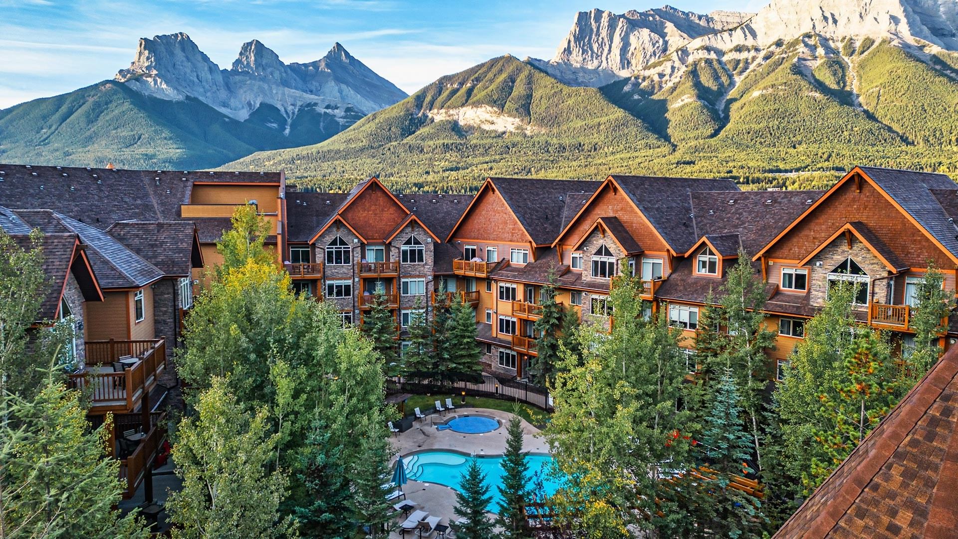 This is a scenic view of a resort with a pool surrounded by trees, set against towering mountain peaks.