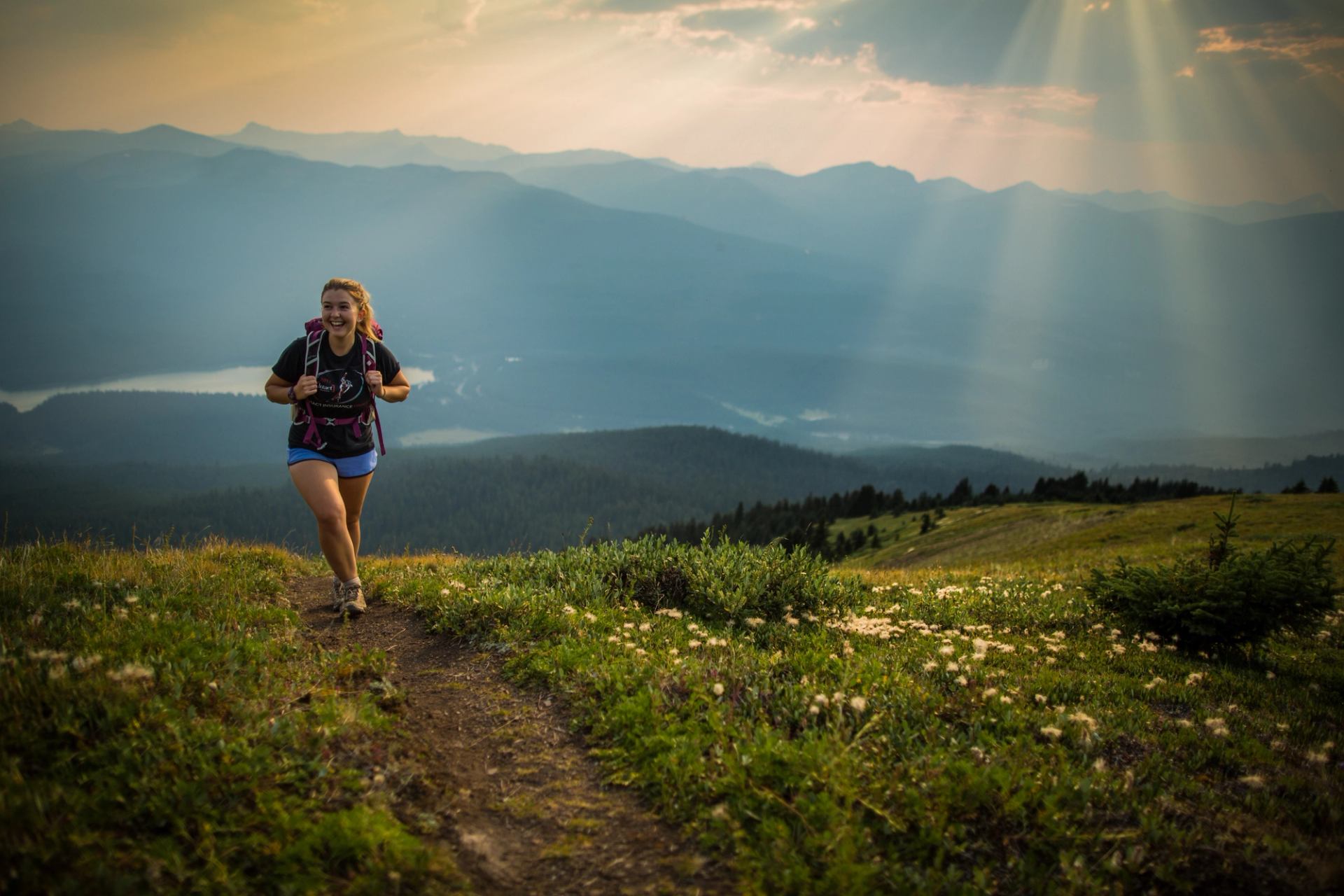 Hiker with backpack runs along mountain trail above valley and distant lake.