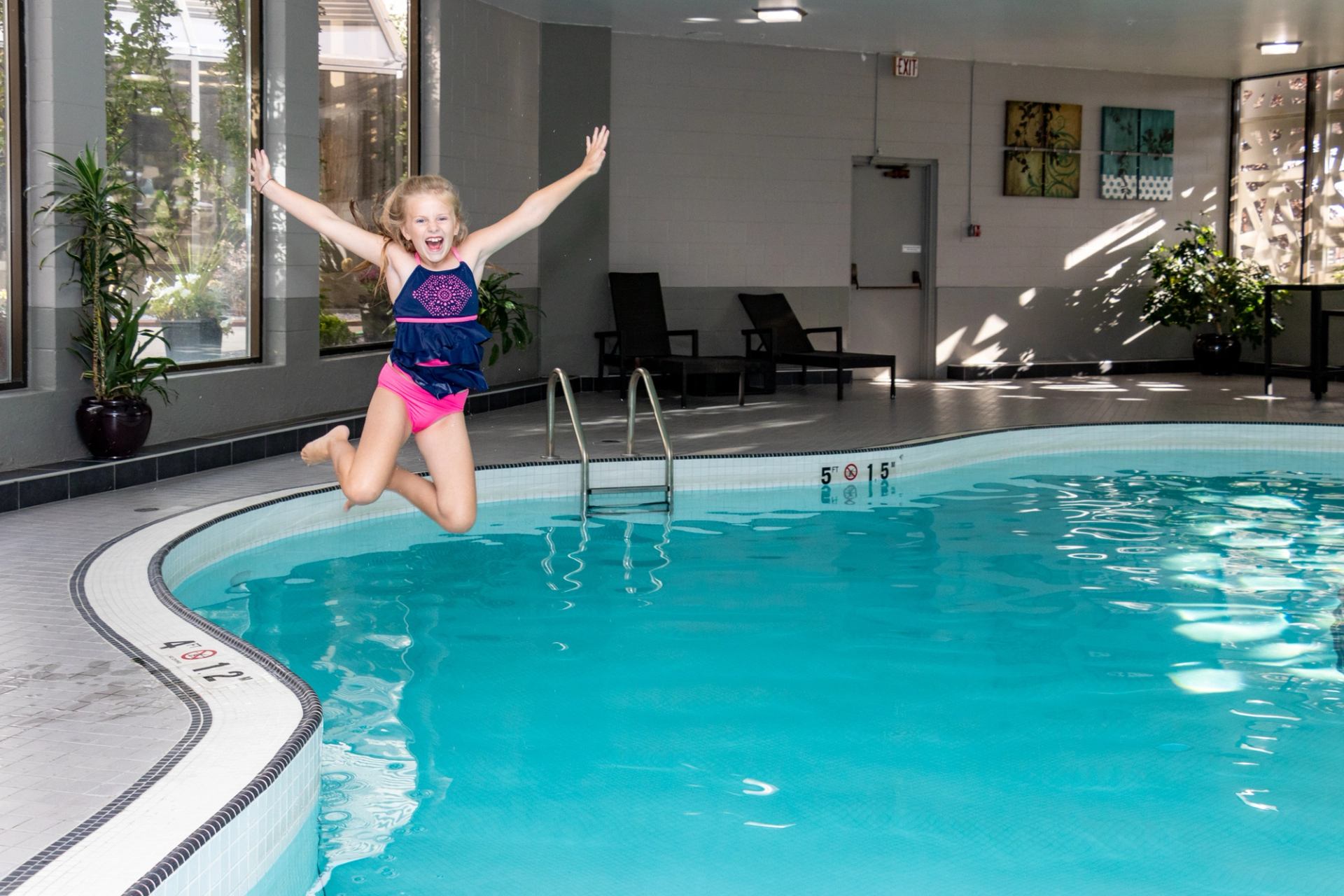 Child in swimsuit jumps into indoor hotel pool near lounge chairs and plants.