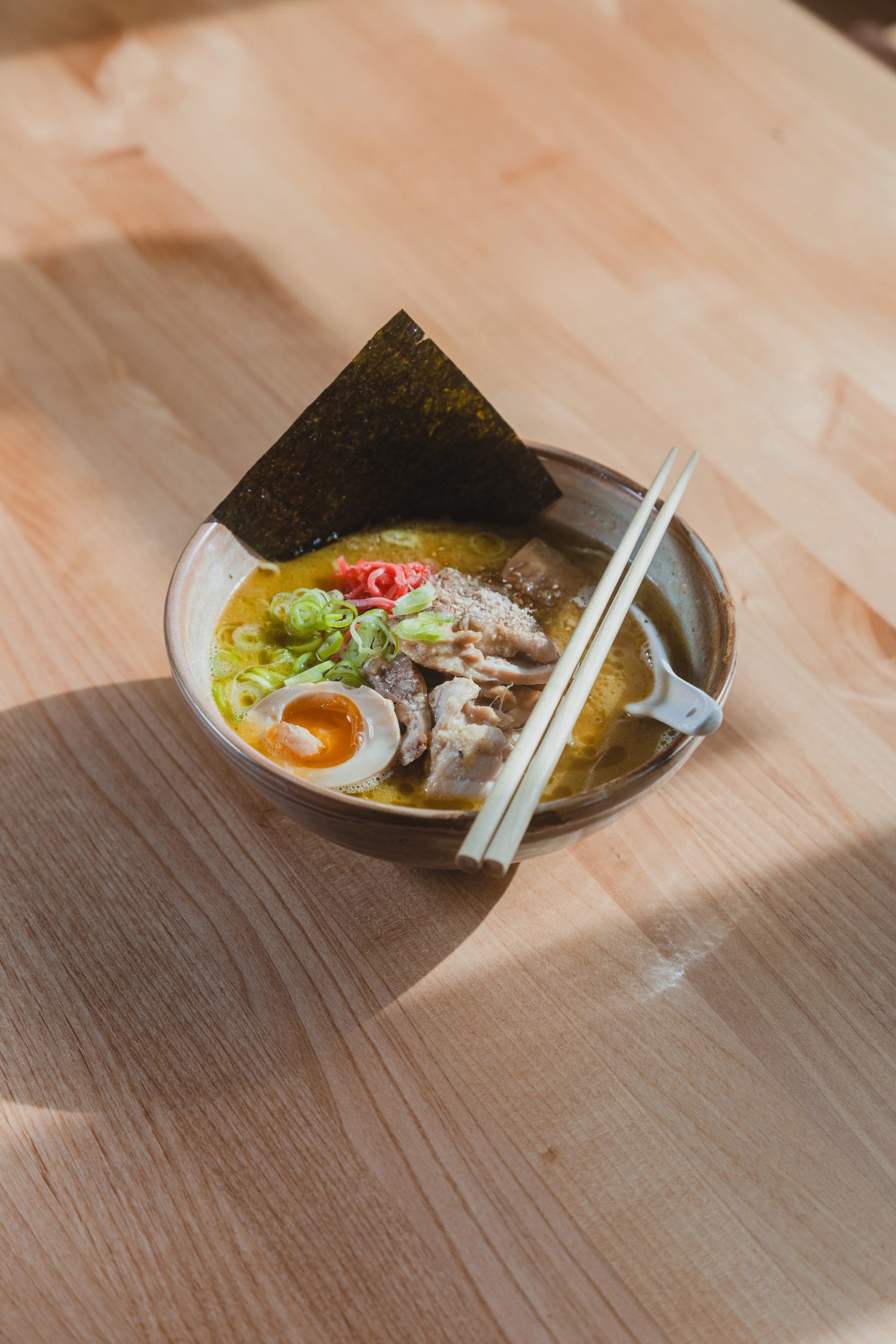 A bowl of ramen with pork, a soft-boiled egg, green onions, red ginger, and nori, with chopsticks resting on top, on a wooden table.