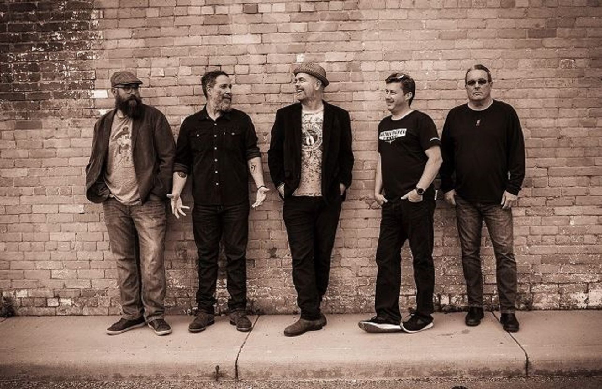 Five musicians standing against a brick wall in a casual lineup.