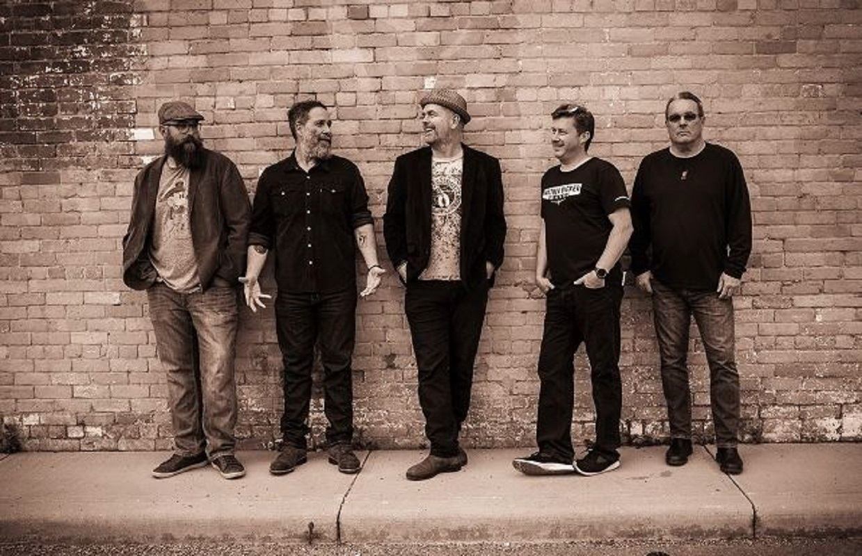 Five musicians standing against a brick wall in a casual lineup.